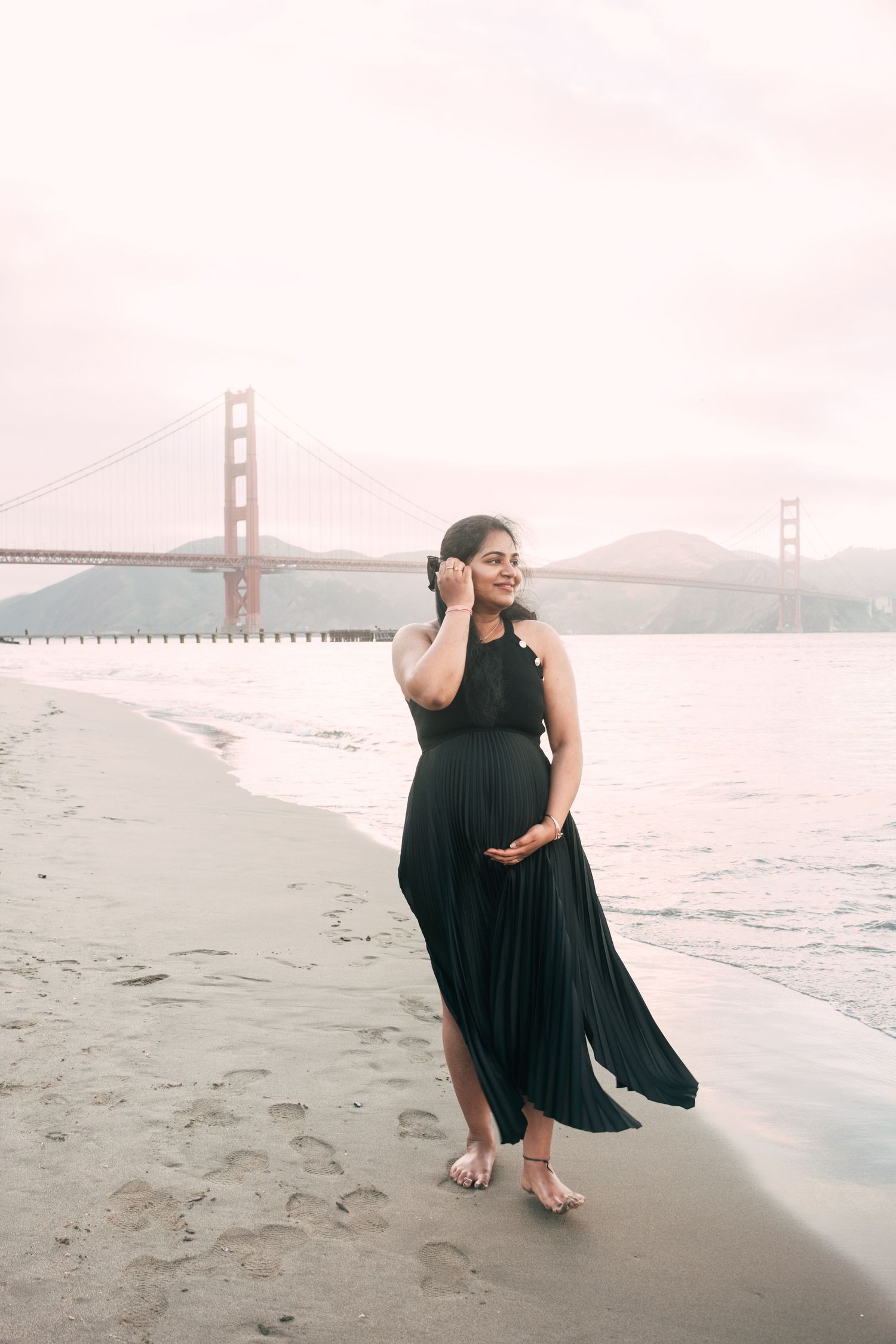 Woman in black dress poses on beach with Golden Gate Bridge in background.