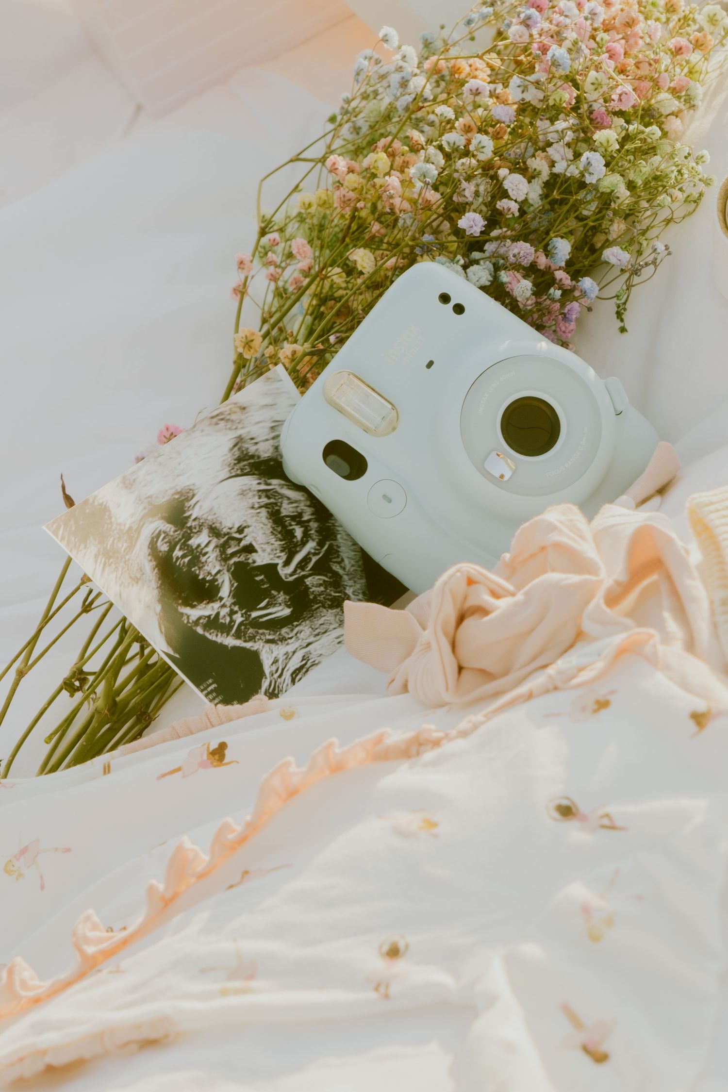 Baby items and delicate flowers arranged on soft white fabric in pastel tones.