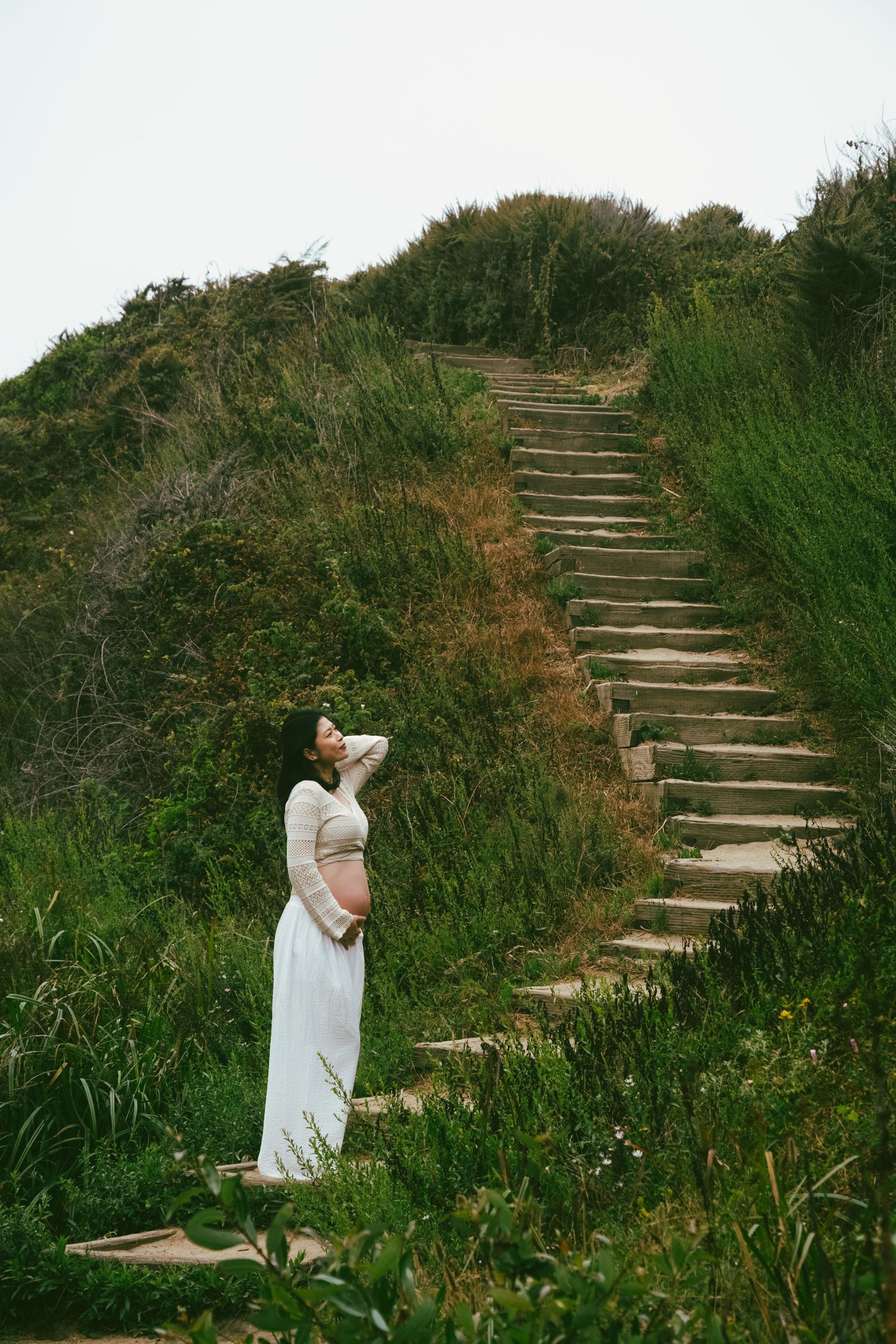 A person in a white dress walks up stone stairs surrounded by lush green vegetation on a hillside.
