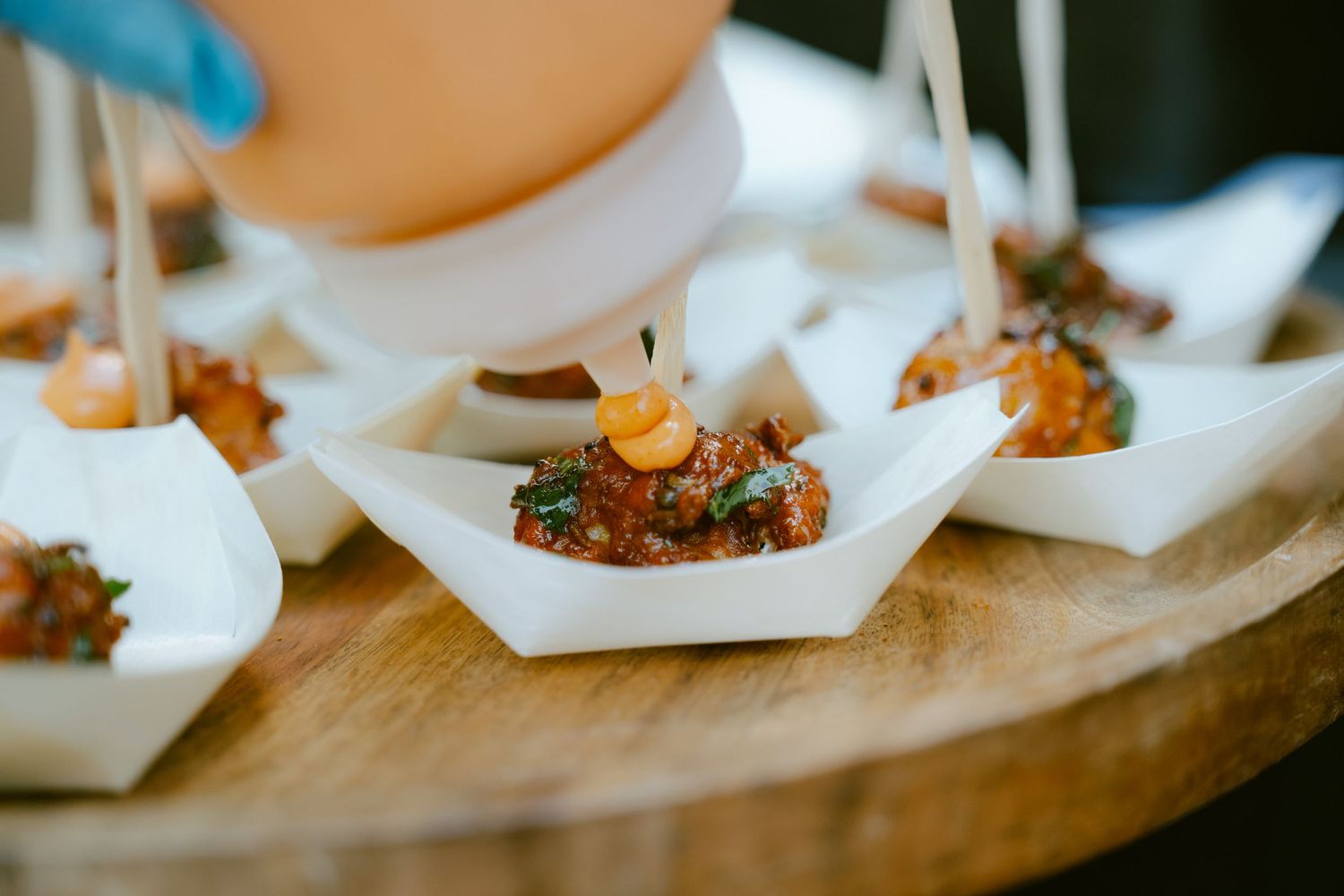 Elegant bite-sized appetizers in white paper boat dishes topped with savory filling, served on a wooden board.