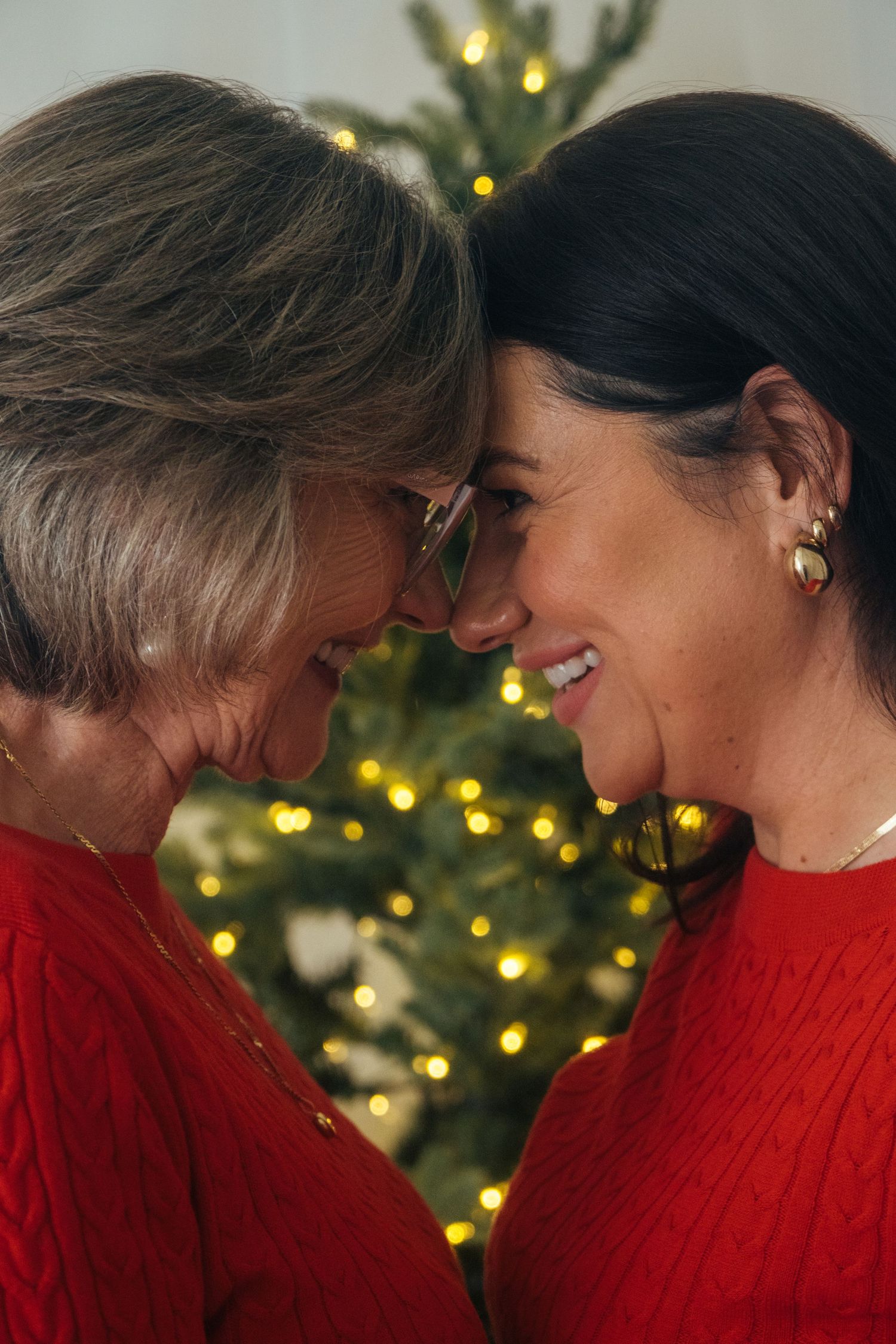Two people in matching red sweaters share intimate moment with foreheads touching near Christmas tree.