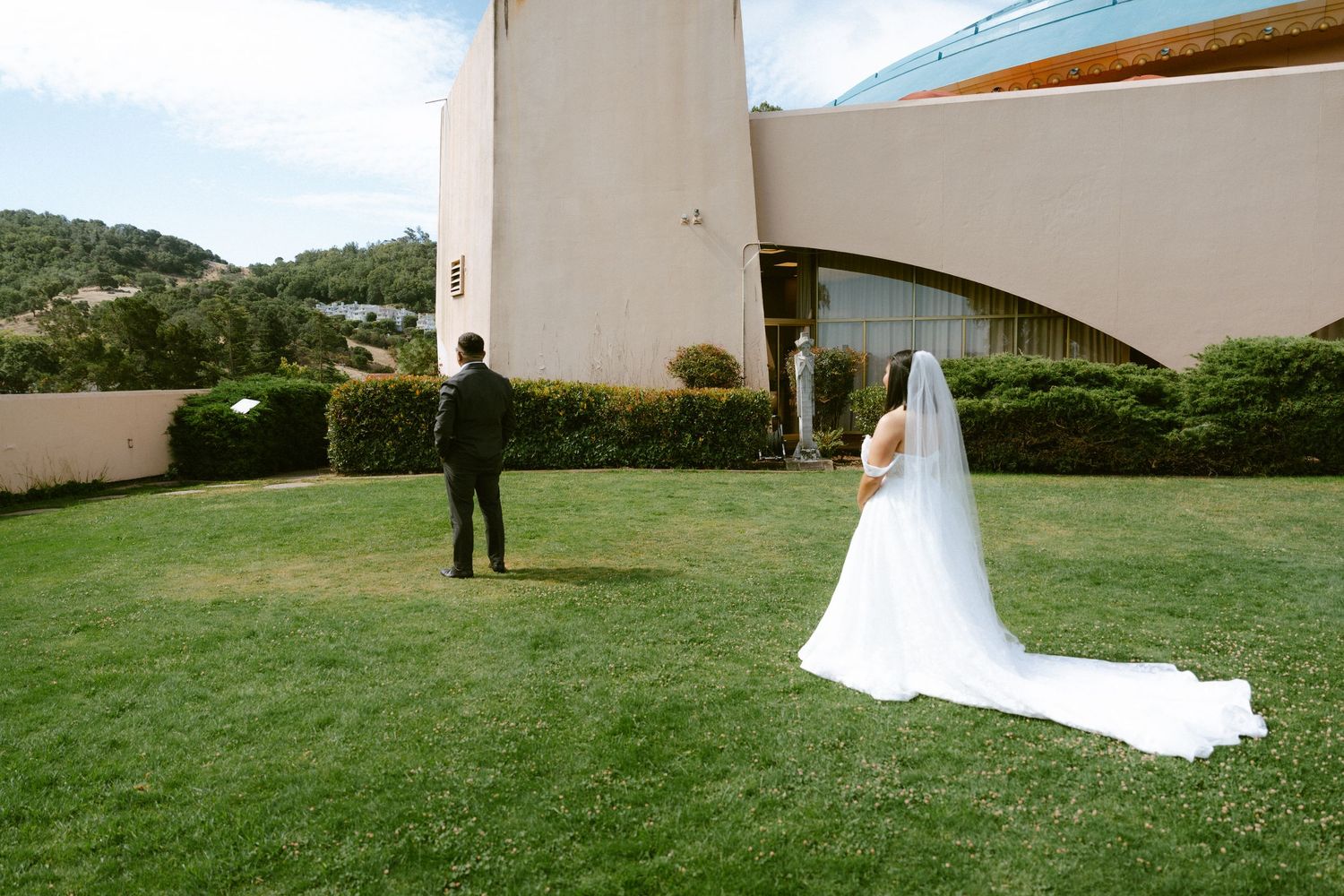 Bride in white gown with veil approaches groom on green lawn beside modern building at outdoor wedding venue.