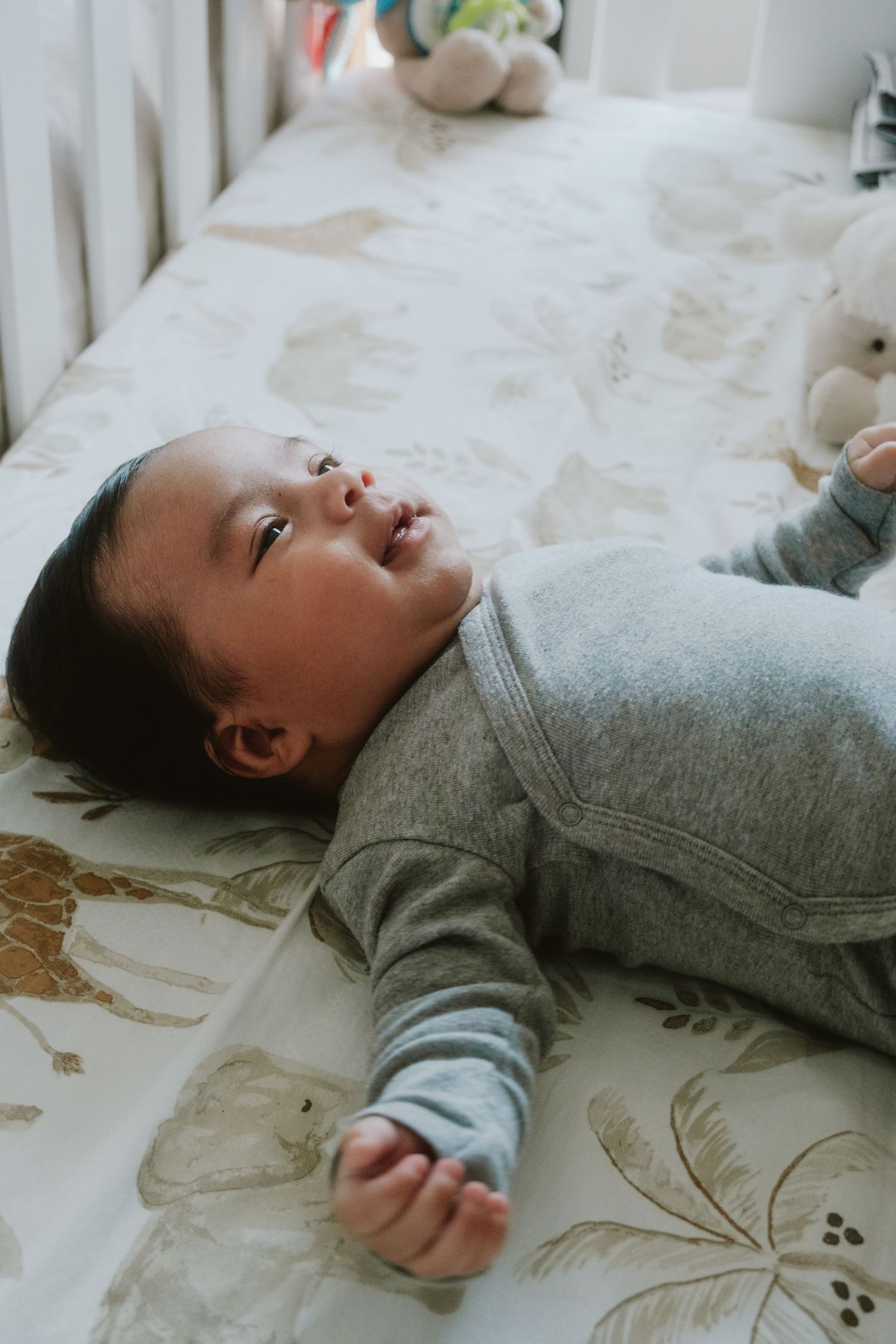 Baby in gray onesie lying in a white crib with animal print sheets and stuffed toys nearby.