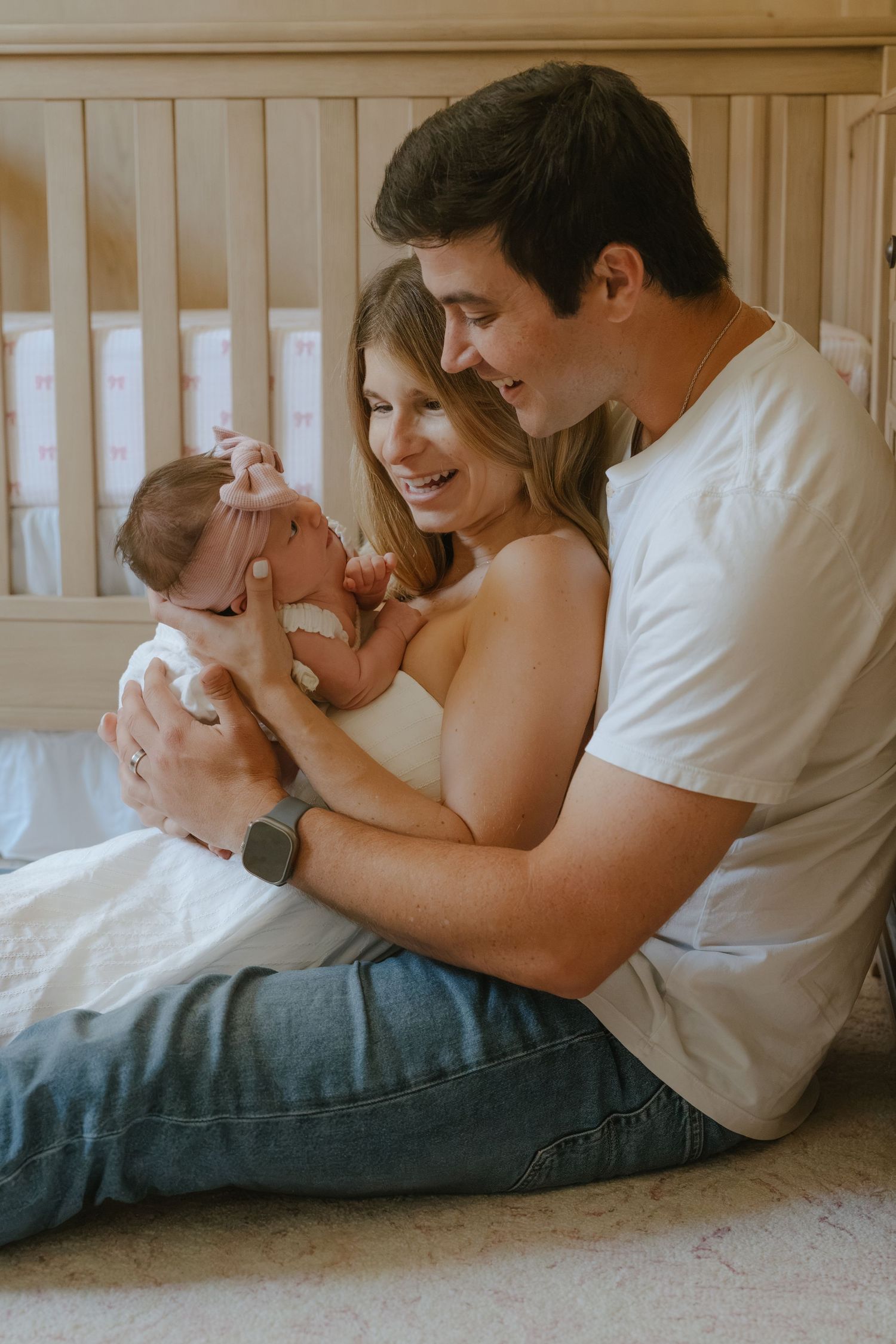 Happy couple sitting on floor holding their newborn baby in a nursery room with wooden crib.