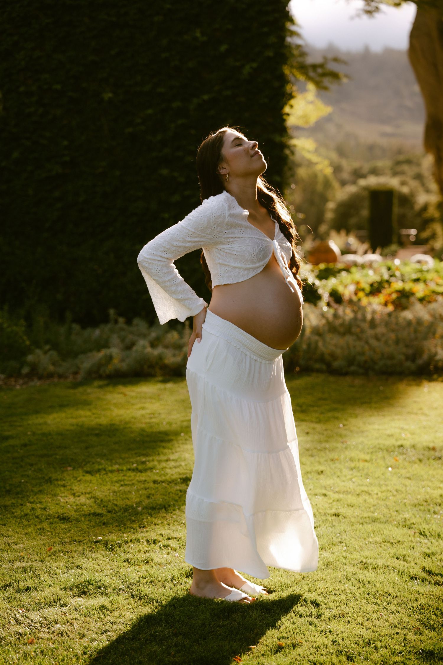 Pregnant woman in white crop top and maxi skirt standing on green lawn in golden sunlight, cradling baby bump.