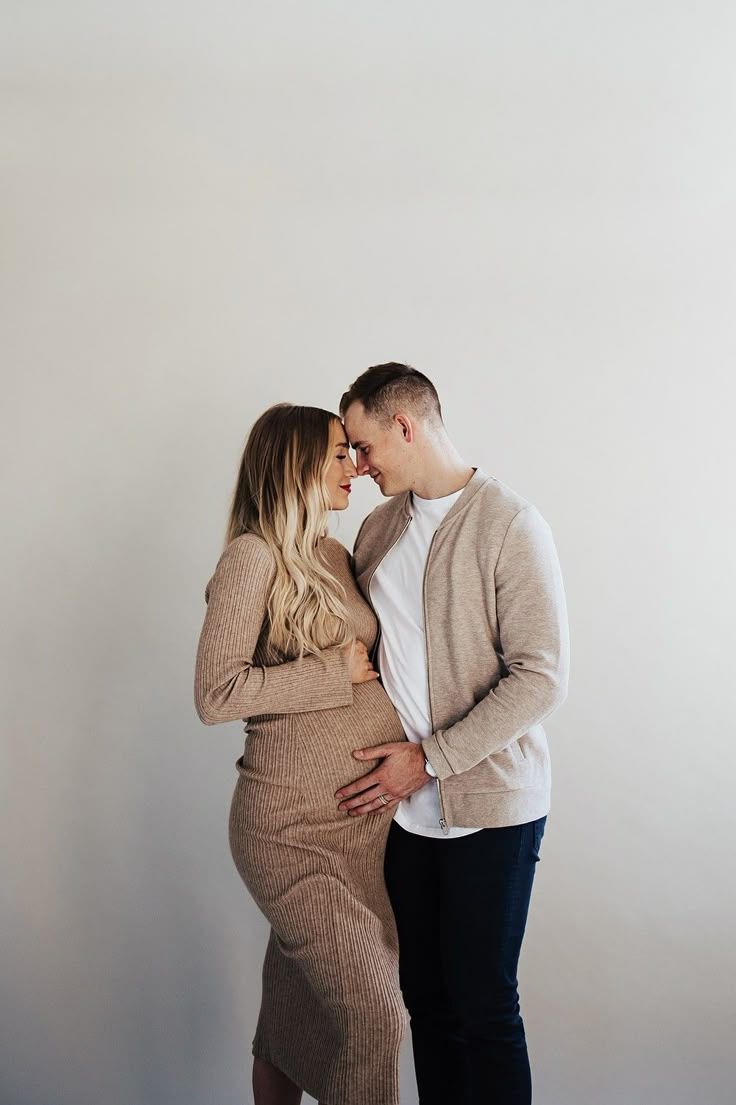 Intimate maternity photoshoot with couple in neutral tones against white wall.