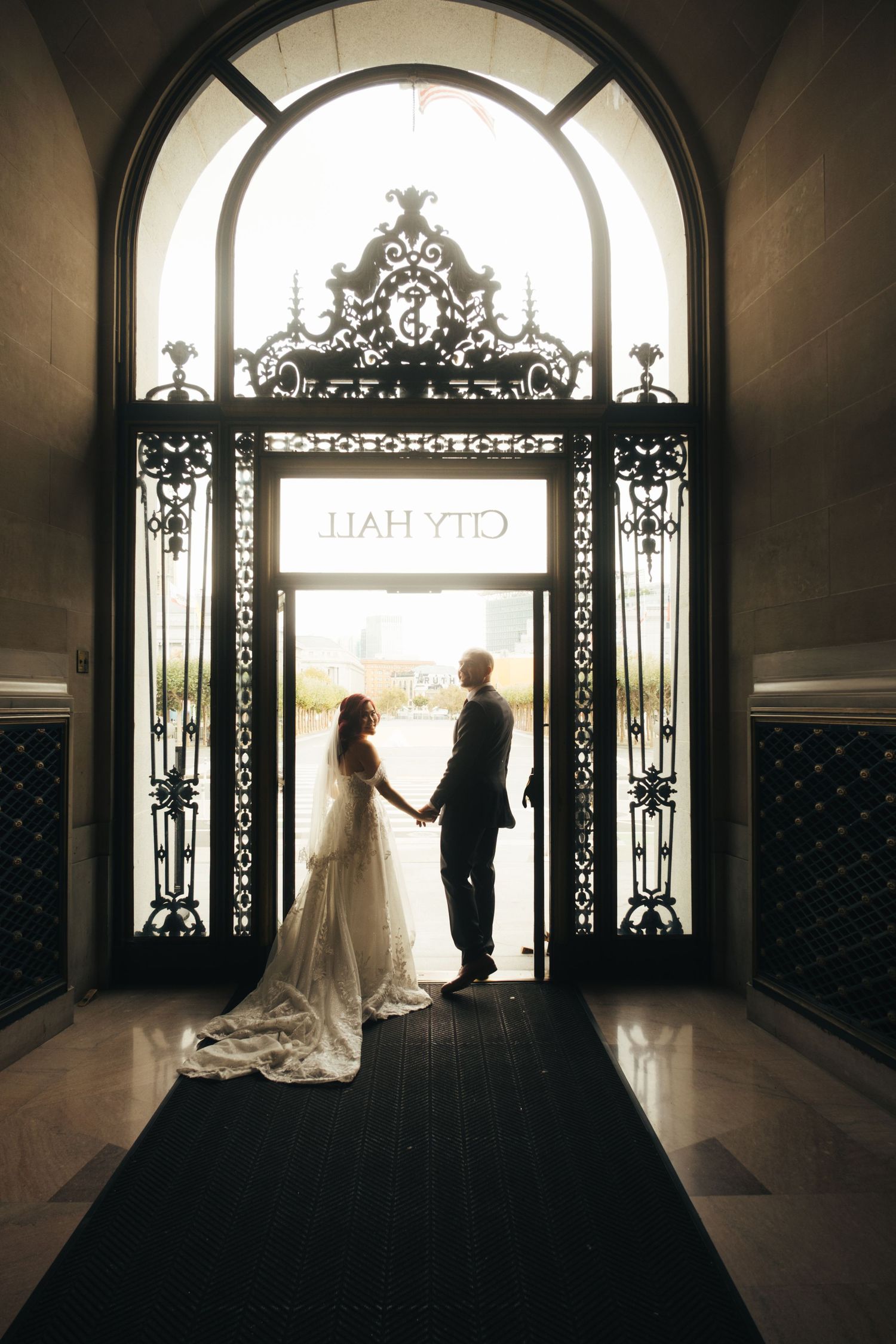Silhouetted couple stands in ornate doorway with City Hall sign above during wedding photoshoot.