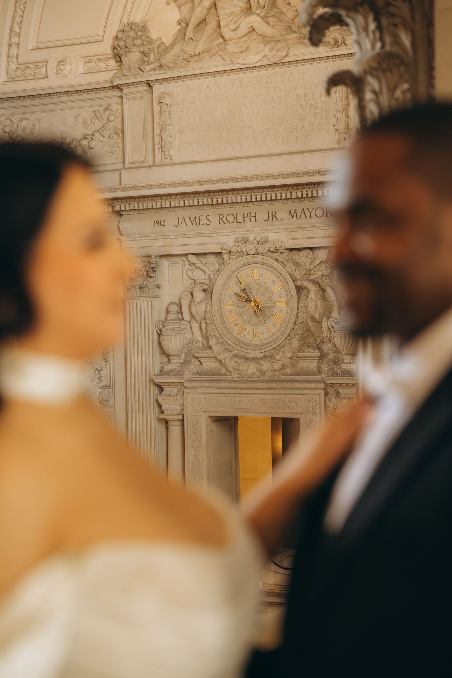 Couple stands before ornate James Rolph Jr. Mayor clock at San Francisco City Hall wedding ceremony.