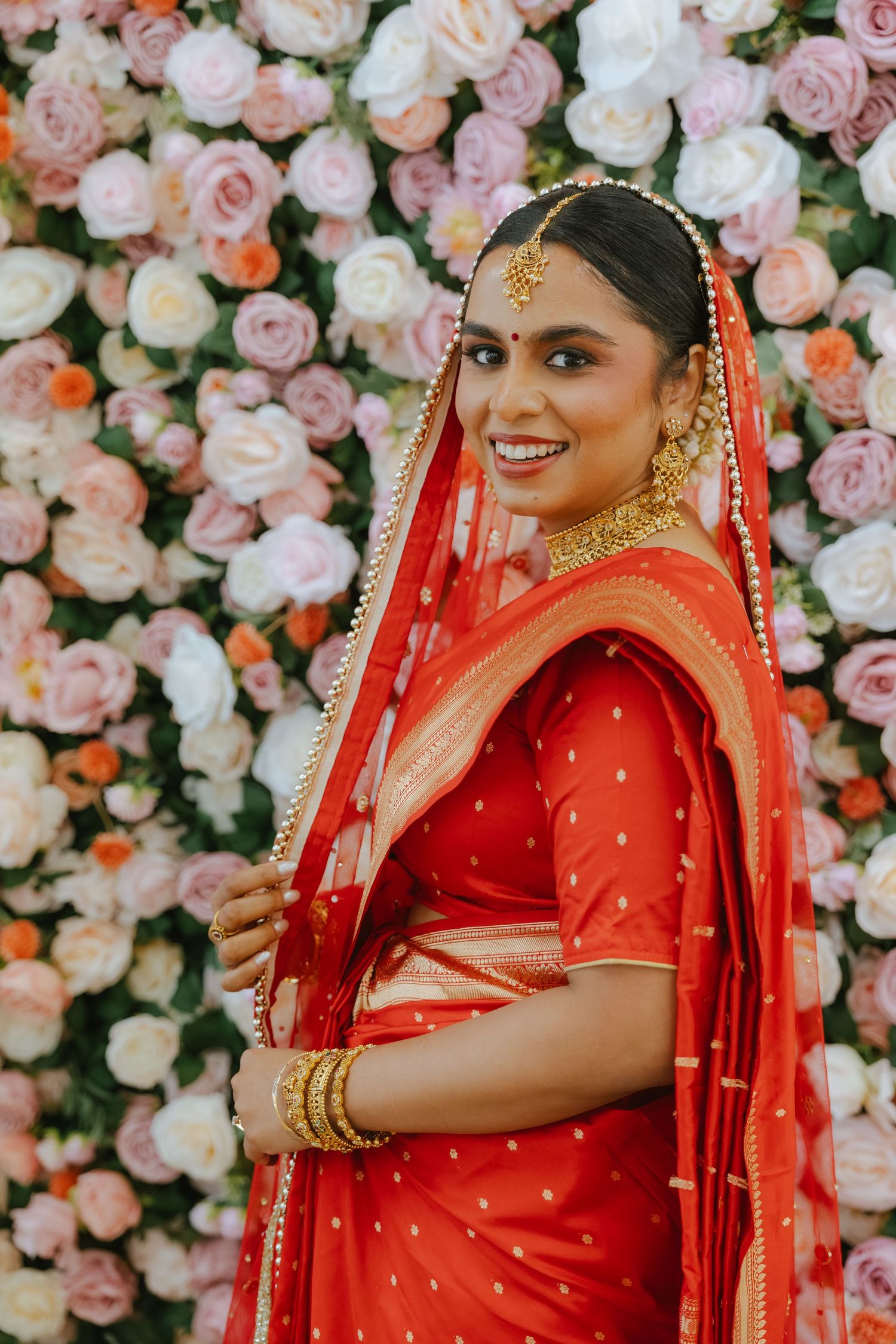 Smiling Indian bride in red saree and gold jewelry poses before a floral wall of pink and white roses.