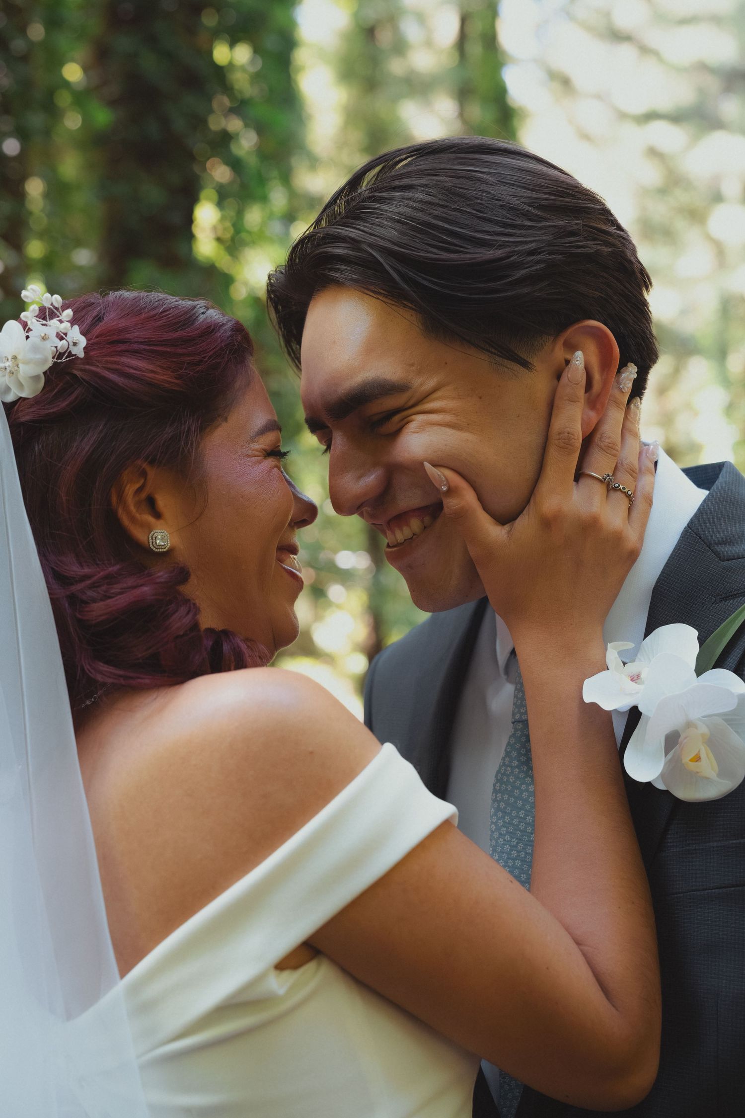 Smiling bride with red hair tenderly holds grooms face as they laugh together outdoors on their wedding day.
