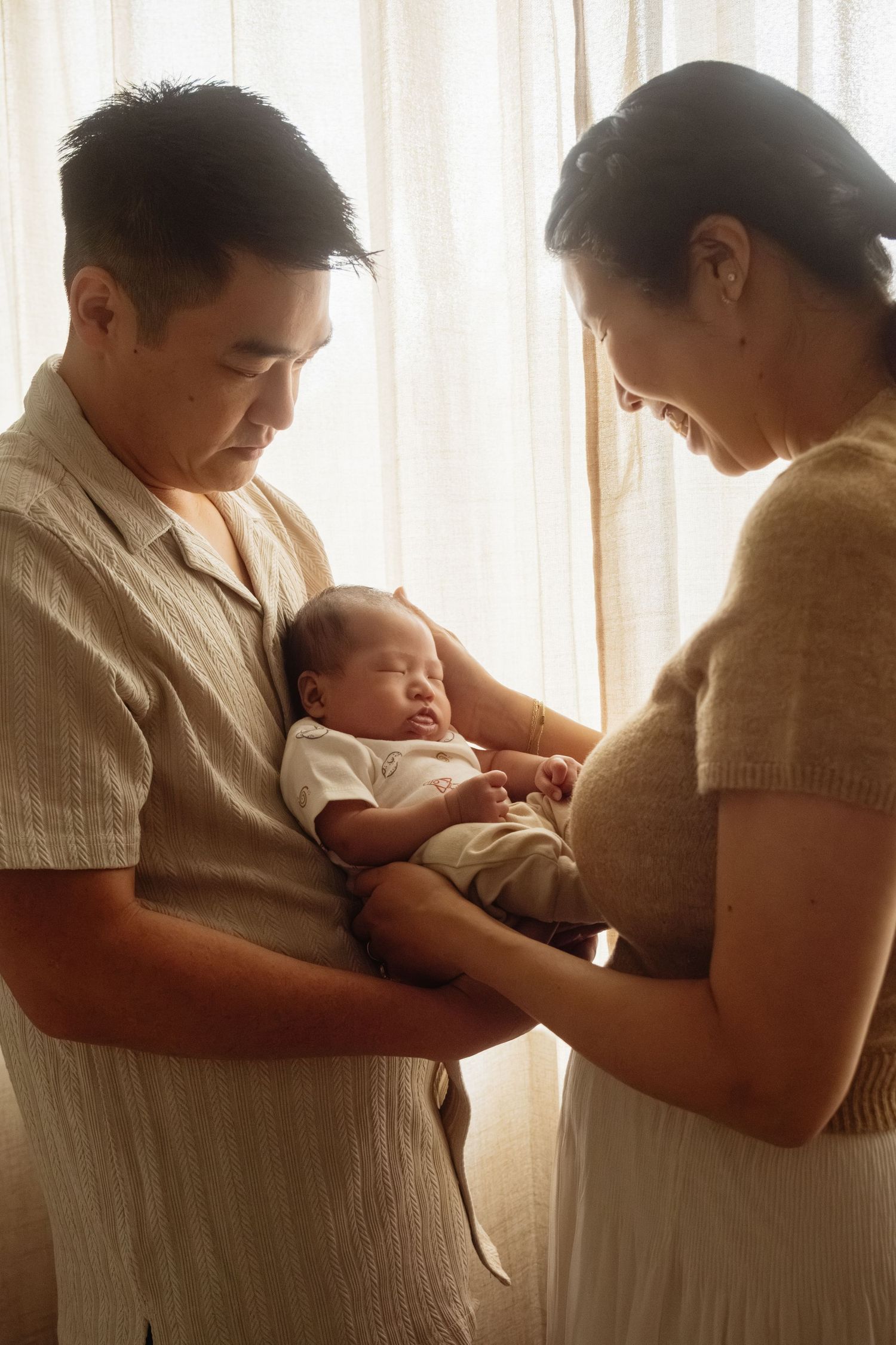 Two parents lovingly gaze at their sleeping newborn baby held in fathers arms near a sunlit window.
