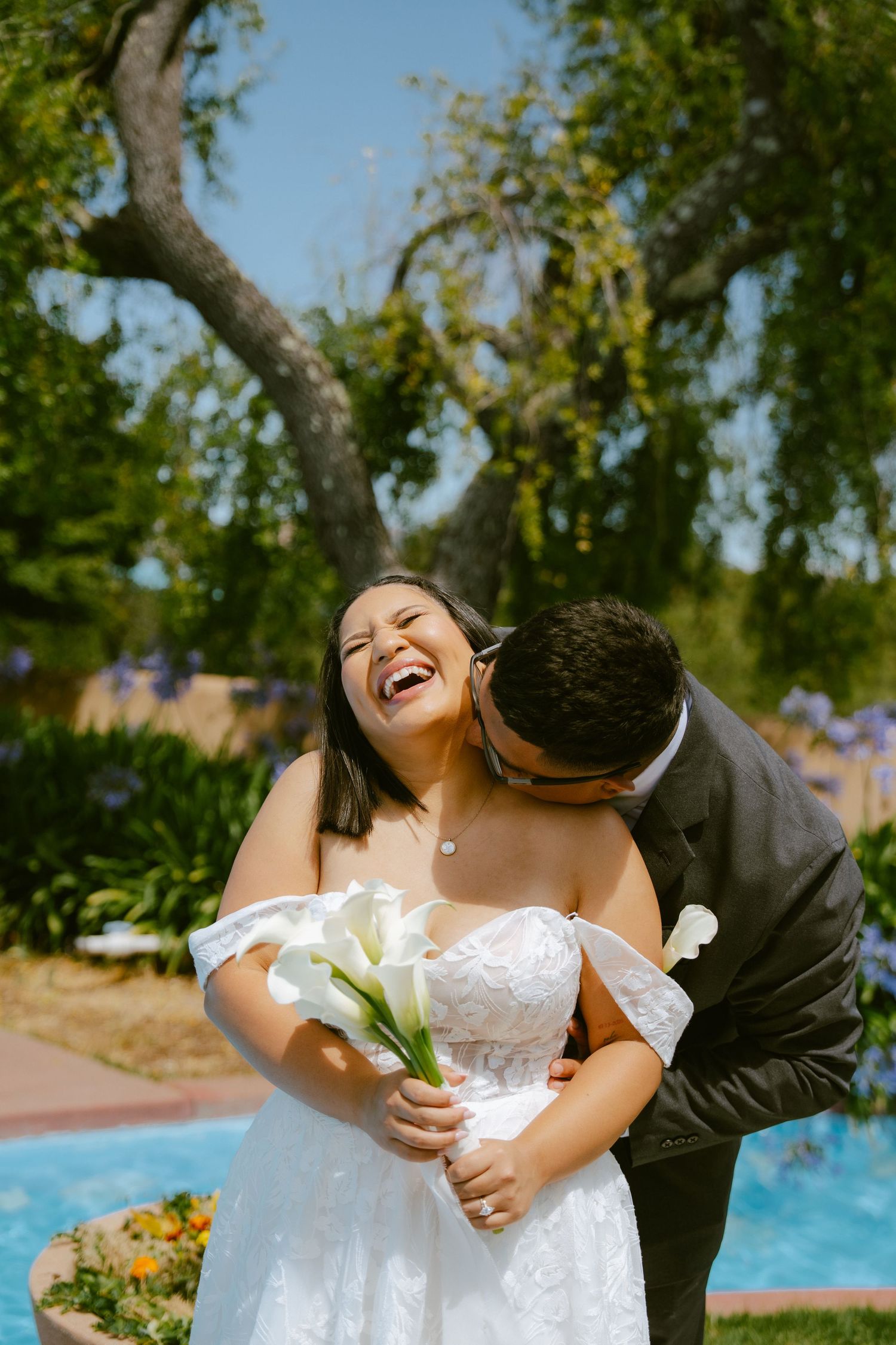 Bride laughing joyfully as groom kisses her cheek outdoors, holding white calla lily bouquet in a lush garden setting.