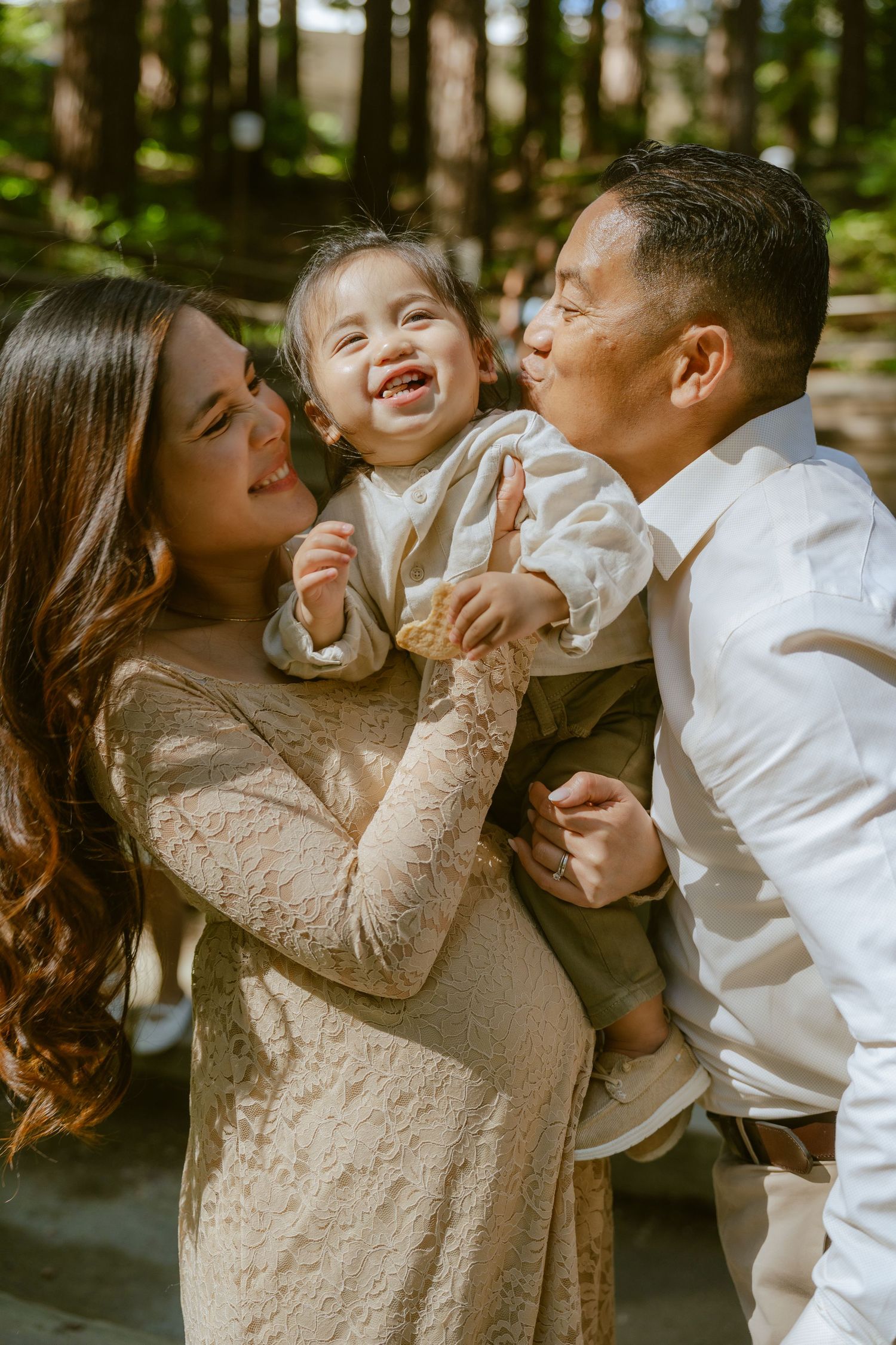 Happy Asian family with mother in lace dress and father kissing laughing baby outdoors in sunny forest.