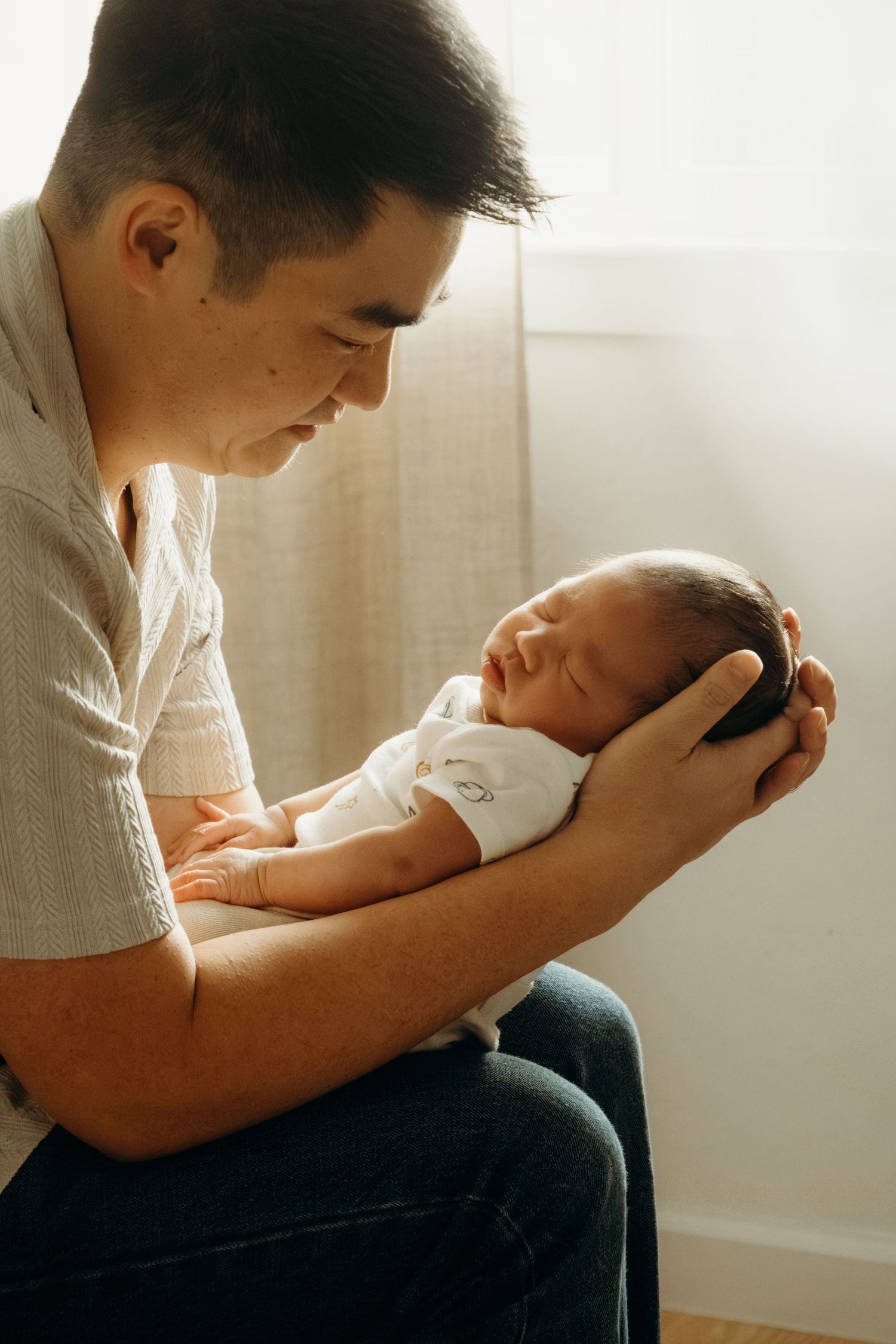 Parent holding and gazing at sleeping newborn baby in gentle window light.