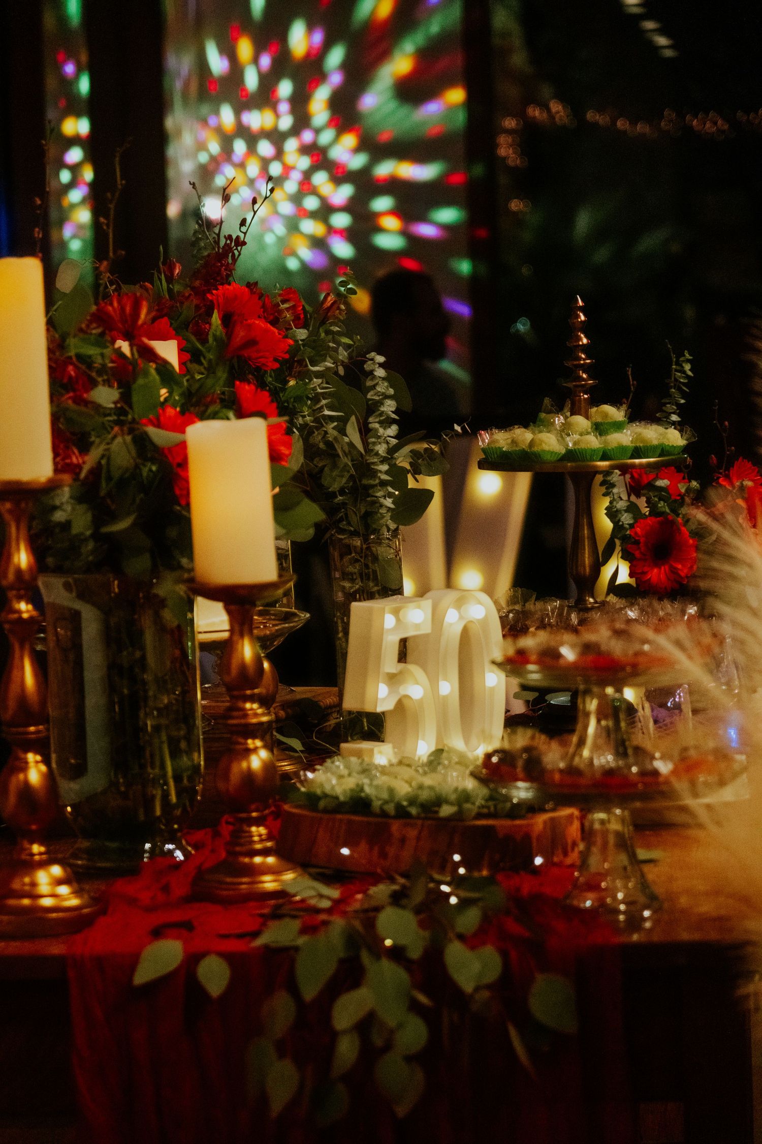 Elegant 50th birthday party table with lit marquee numbers, red roses, white candles, and colorful disco lights in background.