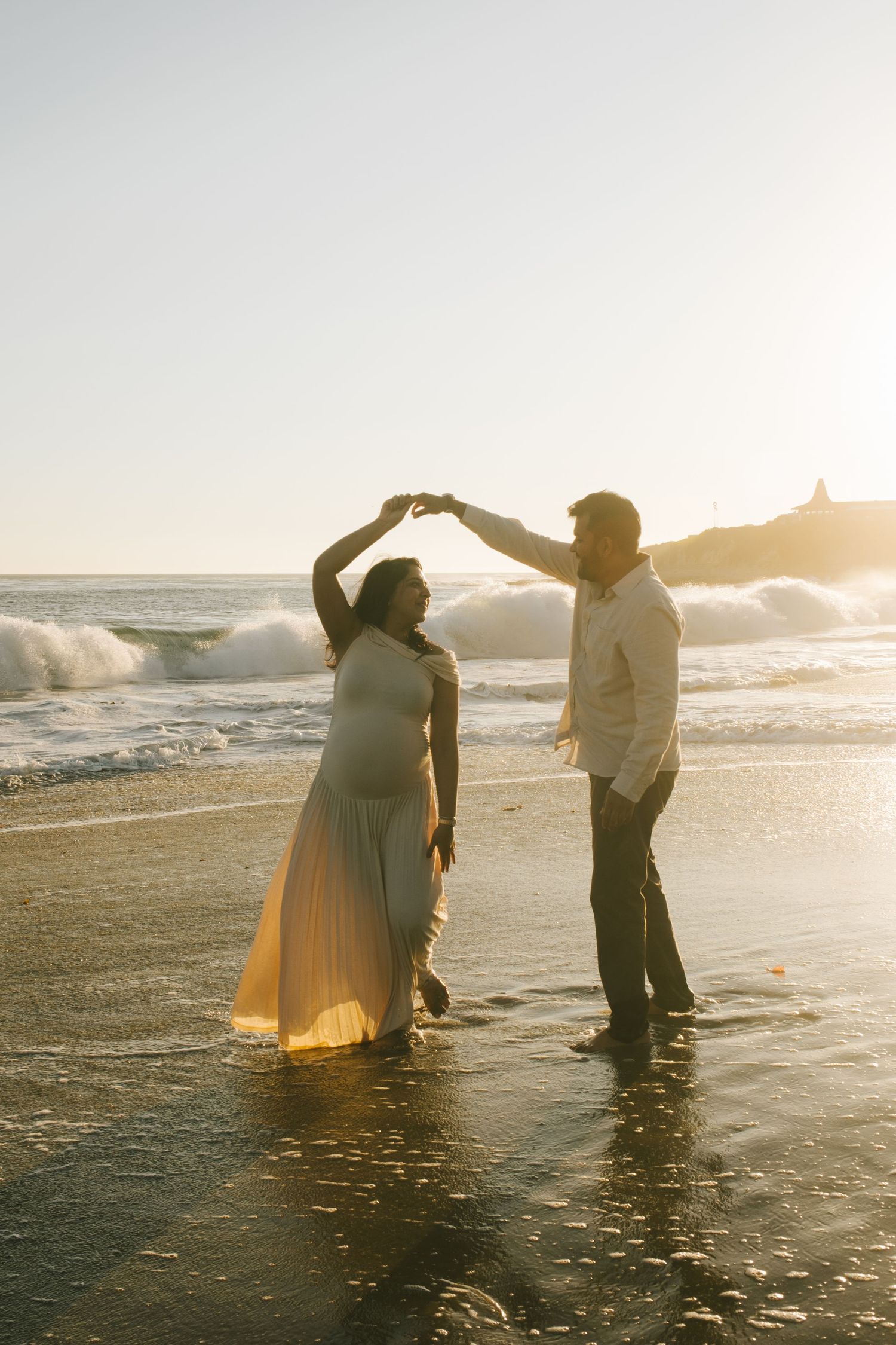 Couple dancing and playing in shallow ocean water during sunset, creating romantic silhouettes.