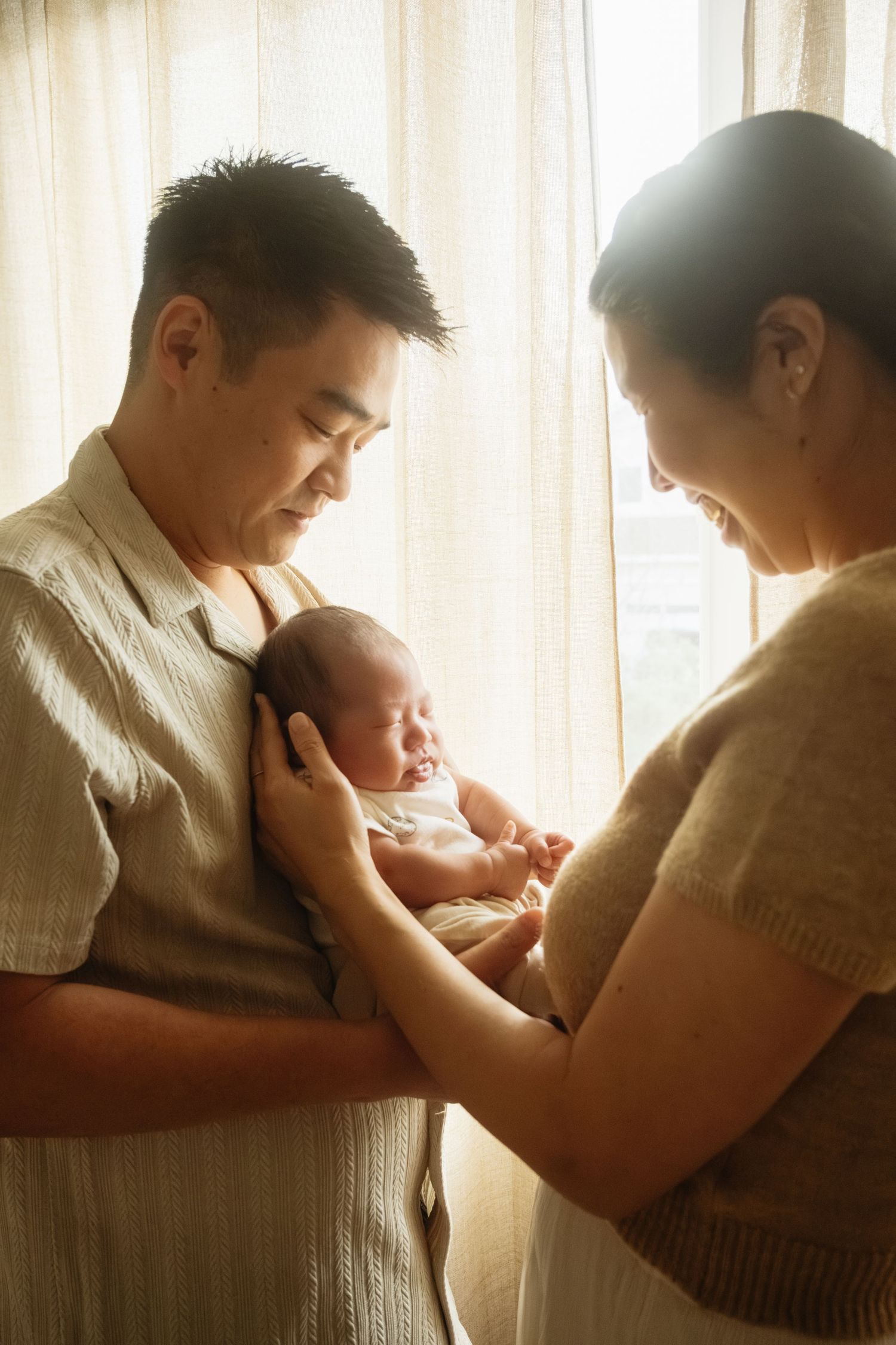New parents share a tender moment with their newborn baby near a sunlit window.