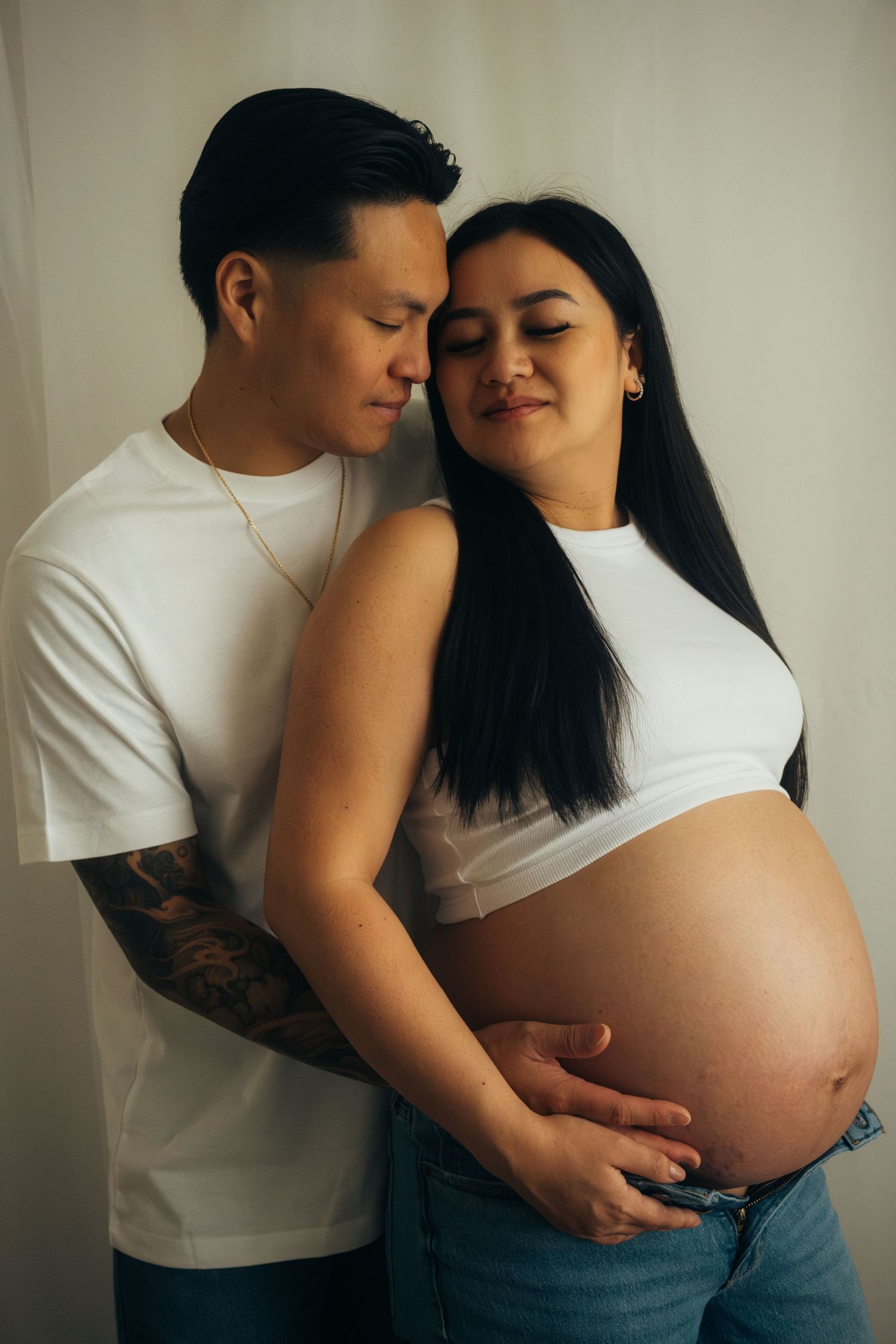 Intimate maternity portrait of couple in white and denim against neutral wall.