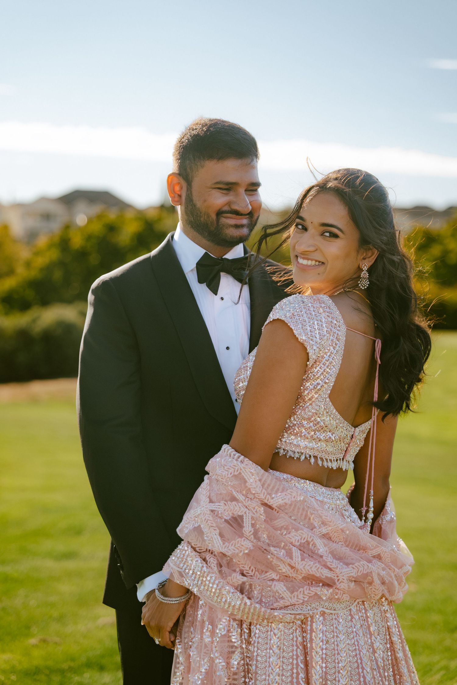 Couple in formal attire, man in tuxedo and woman in embellished pink lehenga, posing outdoors on green lawn.