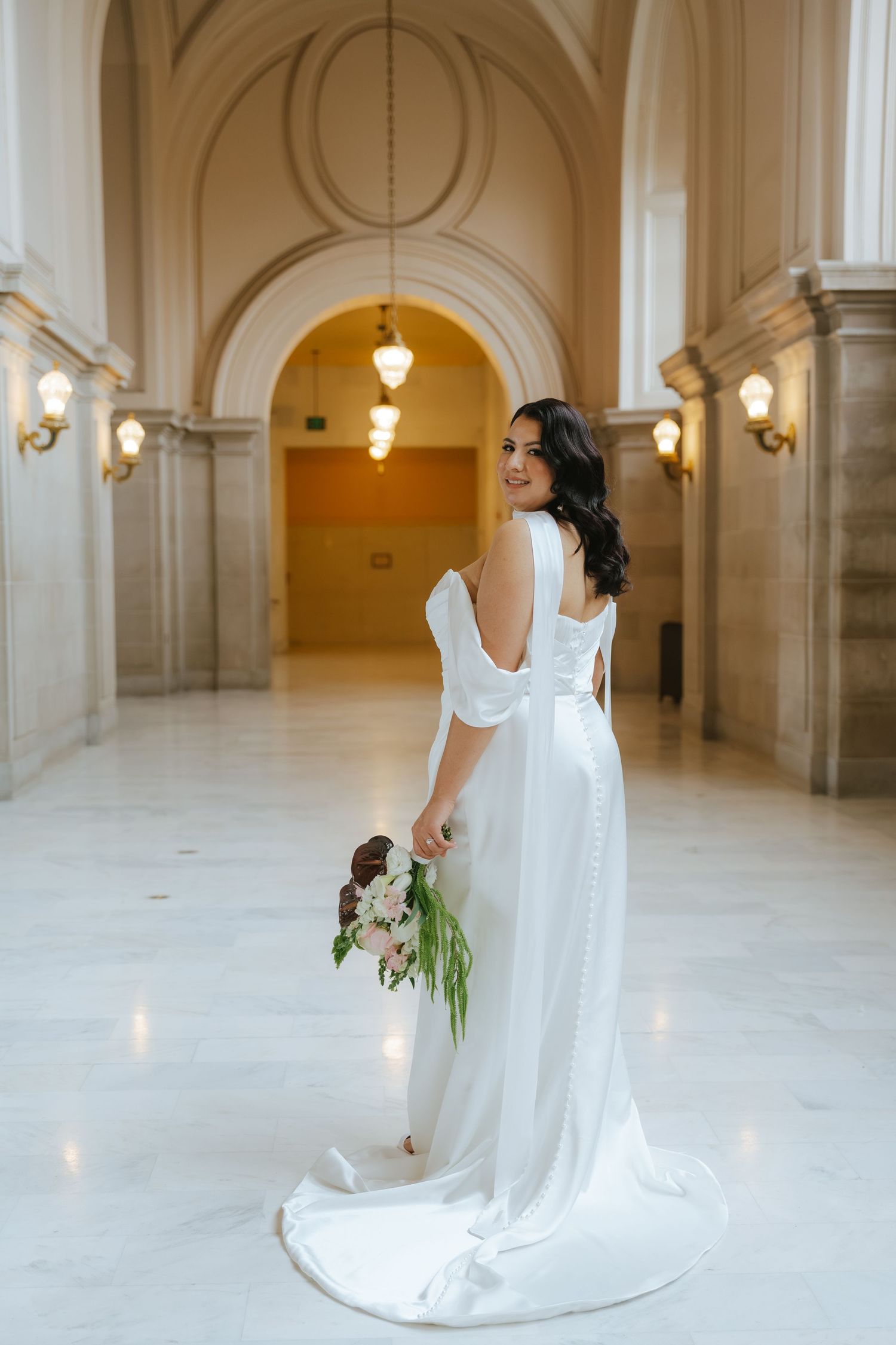 Bride in white gown with open back holds bouquet, smiling over shoulder in elegant marble hall.