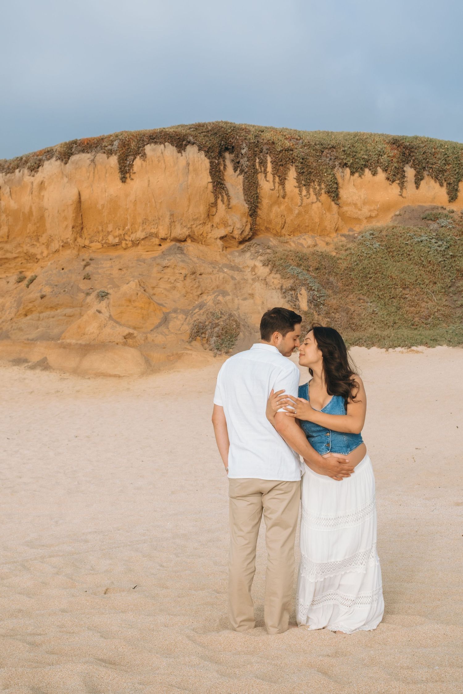 Couple embracing on sandy beach with orange cliffs in background during sunset.