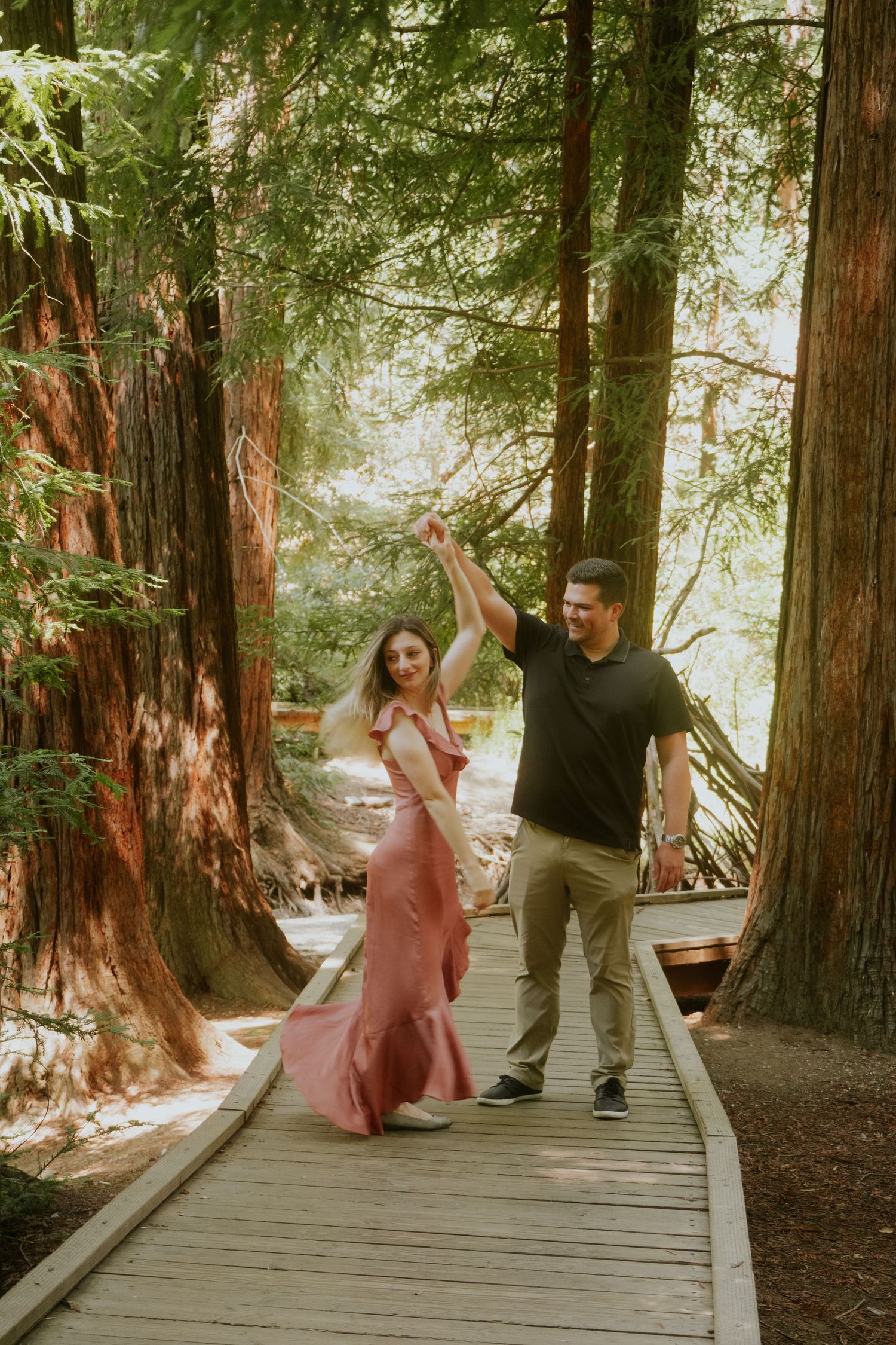 Dancing couple twirls playfully on wooden boardwalk through redwood forest.