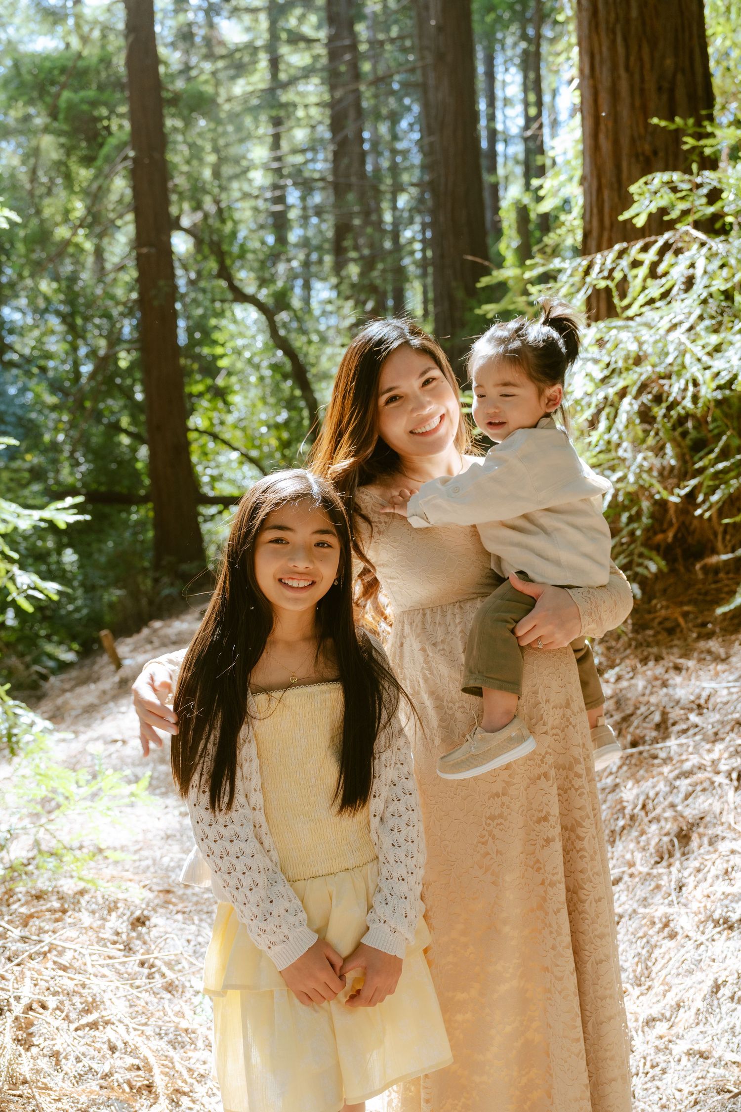 A mother holds her toddler while posing with her daughter in a sunlit forest, all smiling warmly.