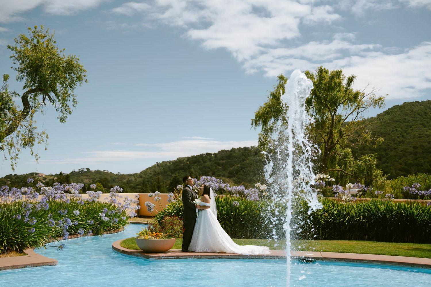 Bride and groom stand by a garden fountain with rolling hills and blue sky at an outdoor wedding venue.