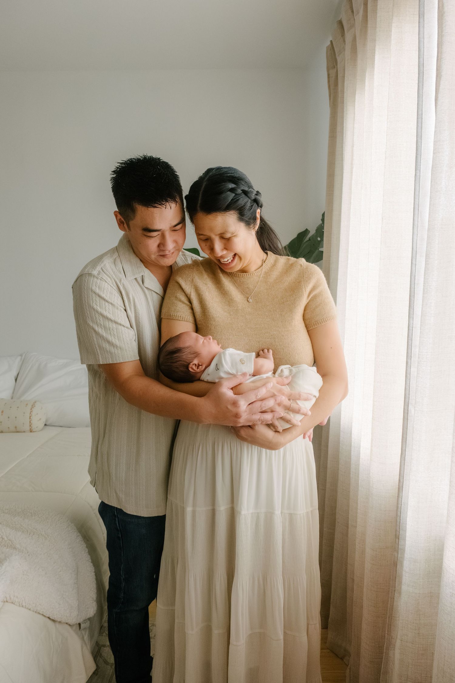 A couple tenderly holds their newborn baby near a bright window in a softly lit bedroom.