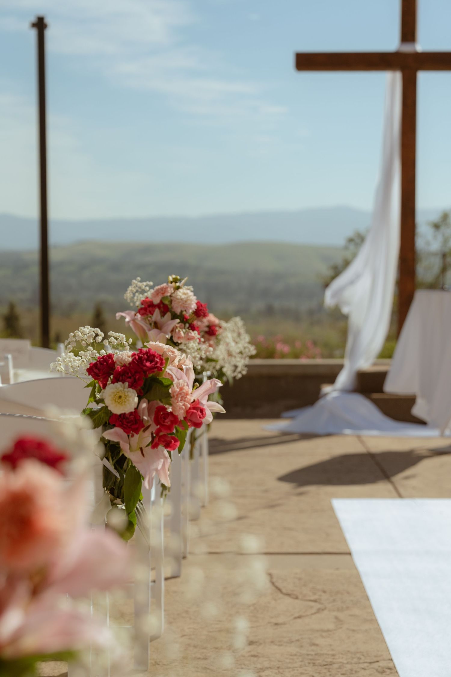 Elegant outdoor wedding ceremony aisle with pink floral arrangements, a wooden cross, and scenic mountain views under blue skies.