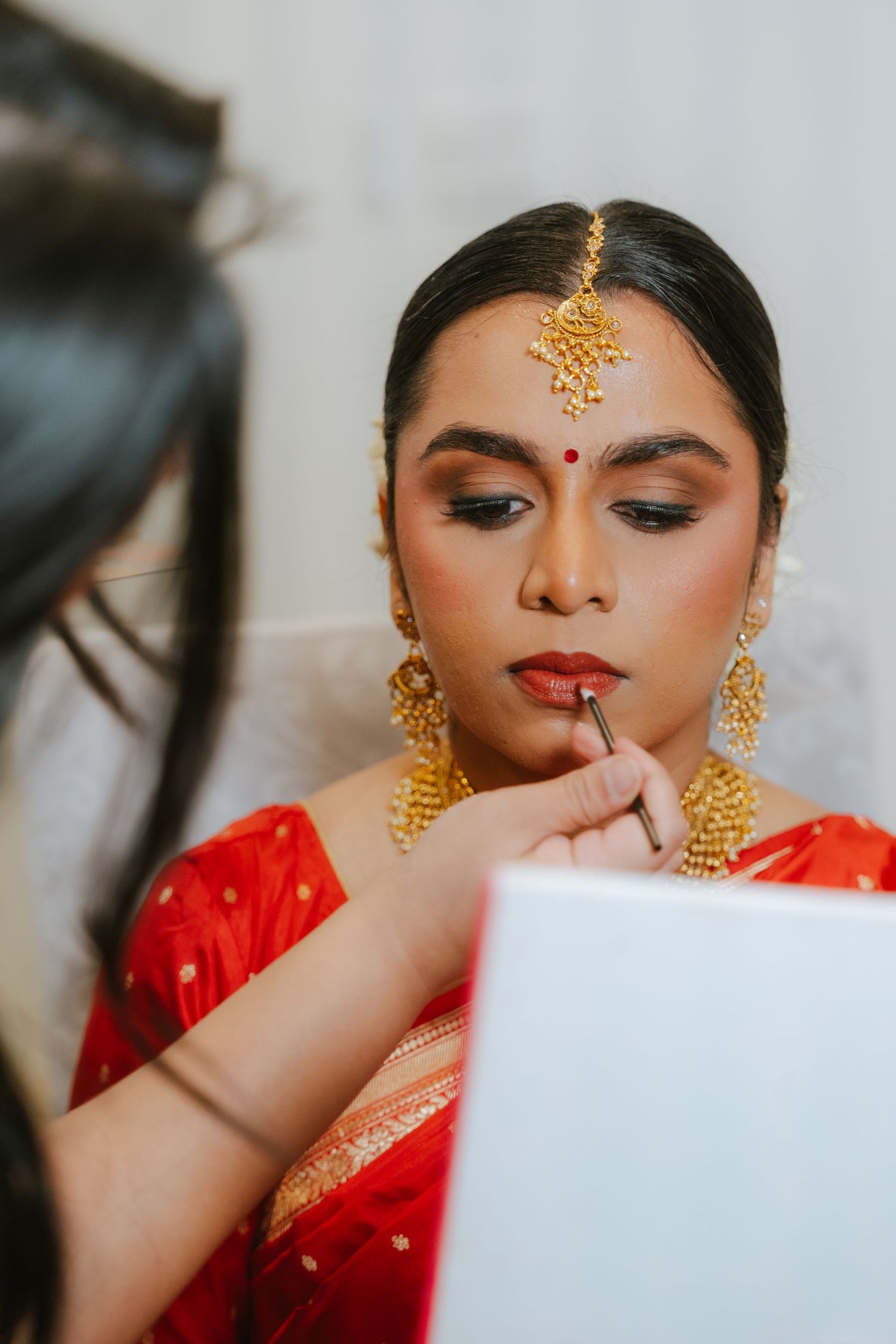 Indian bride in red saree gets lipstick applied by makeup artist, adorned with gold maang tikka and earrings.