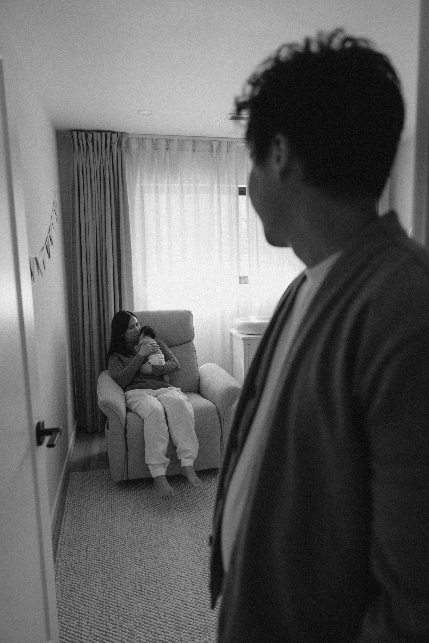 Black and white photo of a mother cradling her newborn in a nursery glider while a man watches from the doorway.