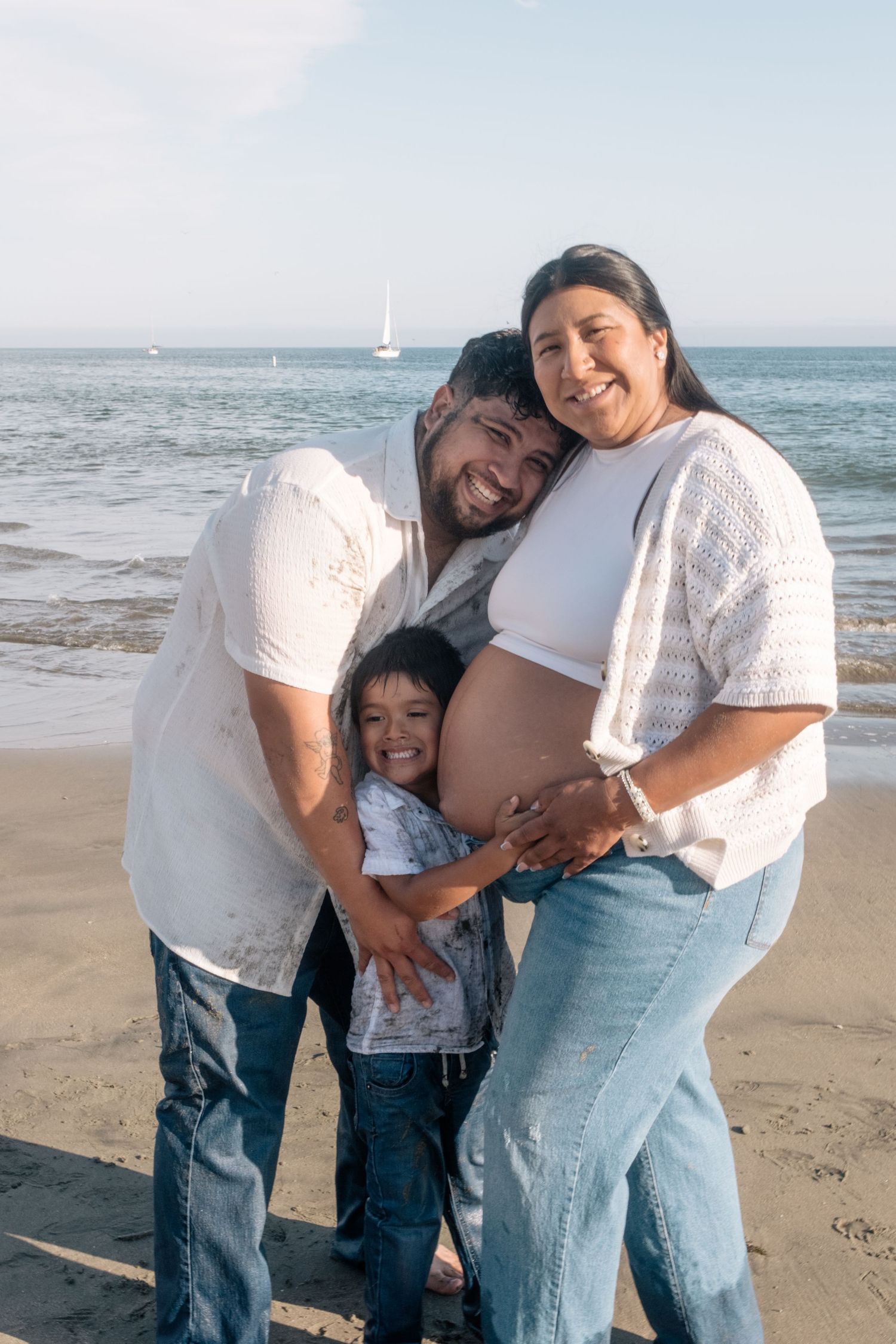 Happy family of three poses on a sunny beach, with pregnant mother, smiling father, and young child hugging her baby bump.