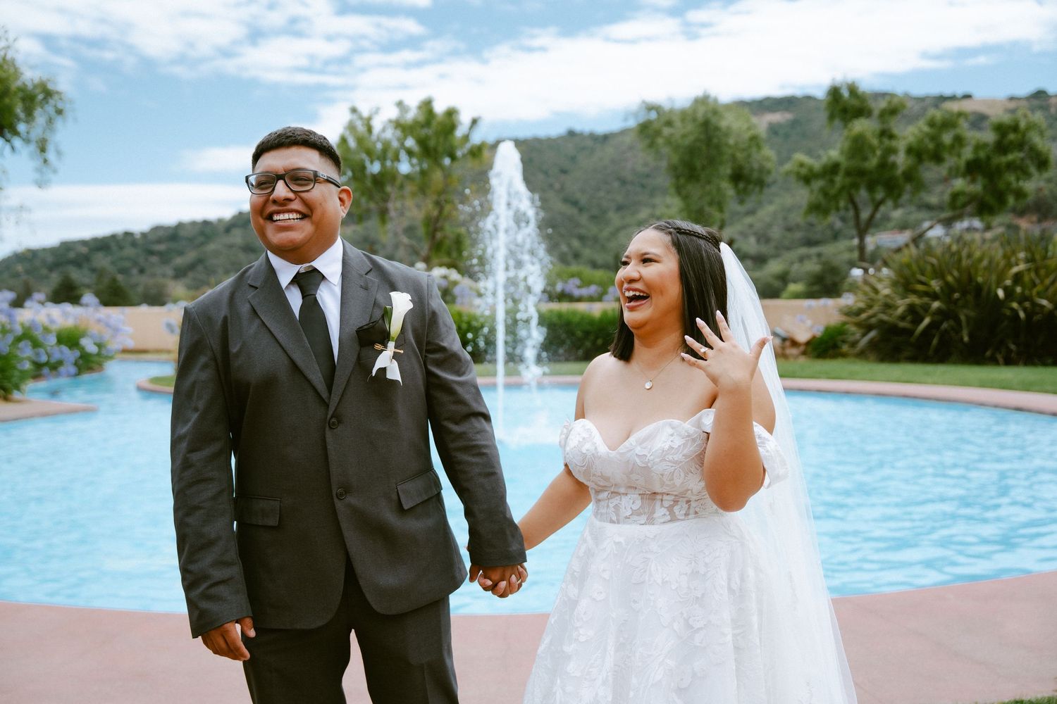 Happy newlyweds holding hands and smiling in front of a fountain, bride in lace gown and groom in gray suit.