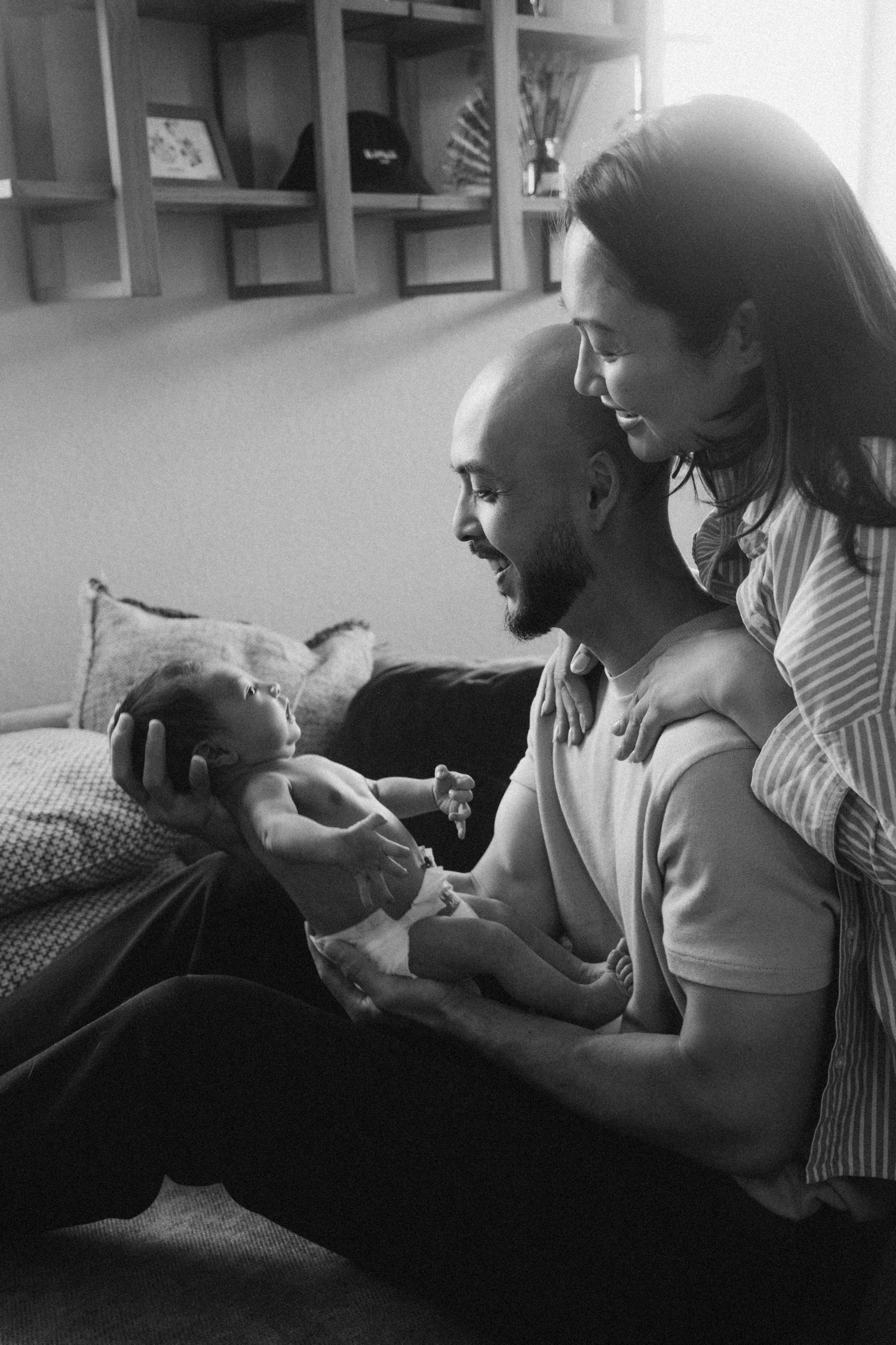 Black and white photo of smiling parents sitting together admiring their newborn baby at home.