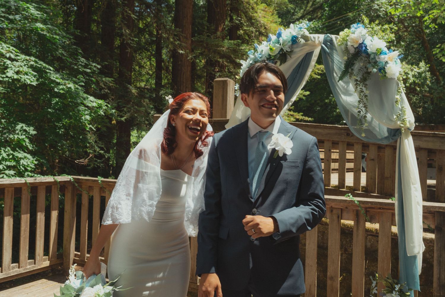 Joyful bride and groom laughing under a blue floral arch in a forest wedding ceremony.