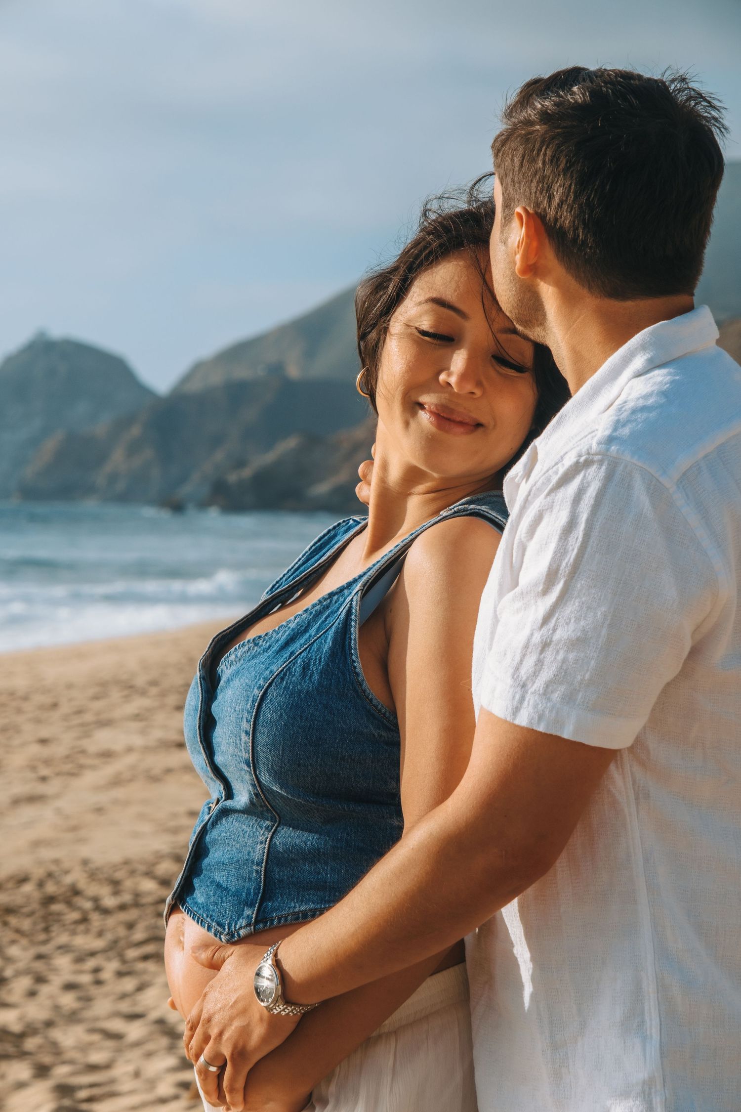 A couple embraces tenderly on a sandy beach with coastal cliffs in the background.