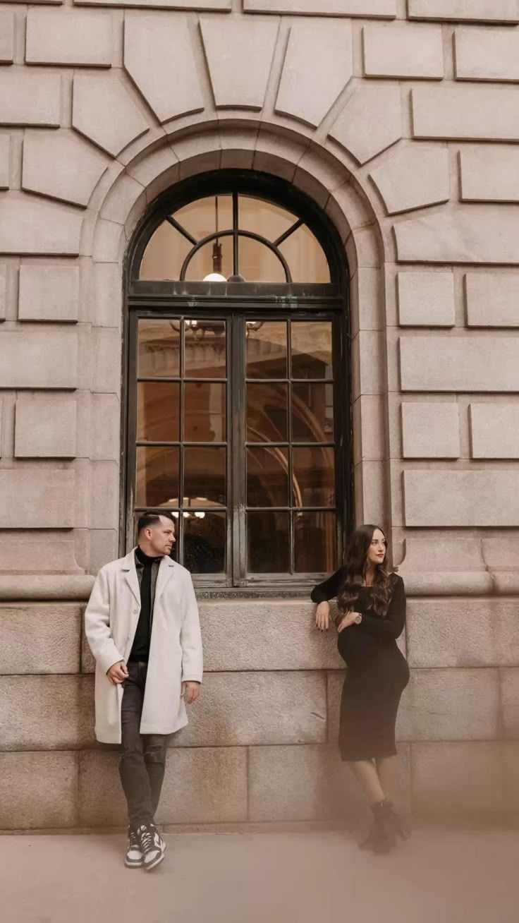 Couple poses playfully in front of arched windows on a classic stone building facade.