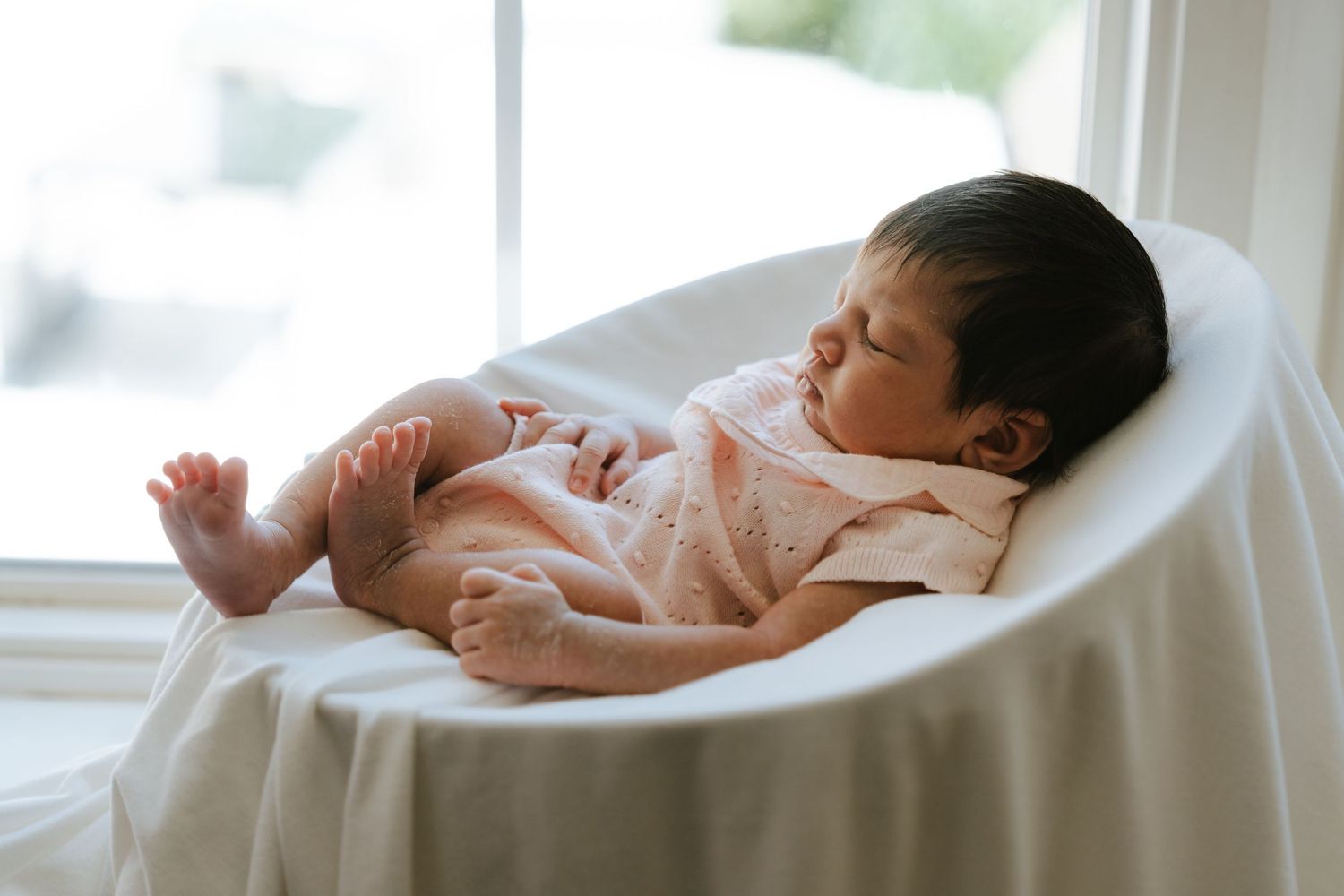 Peaceful newborn baby sleeping in a white round chair near a bright window, wearing a pink onesie.