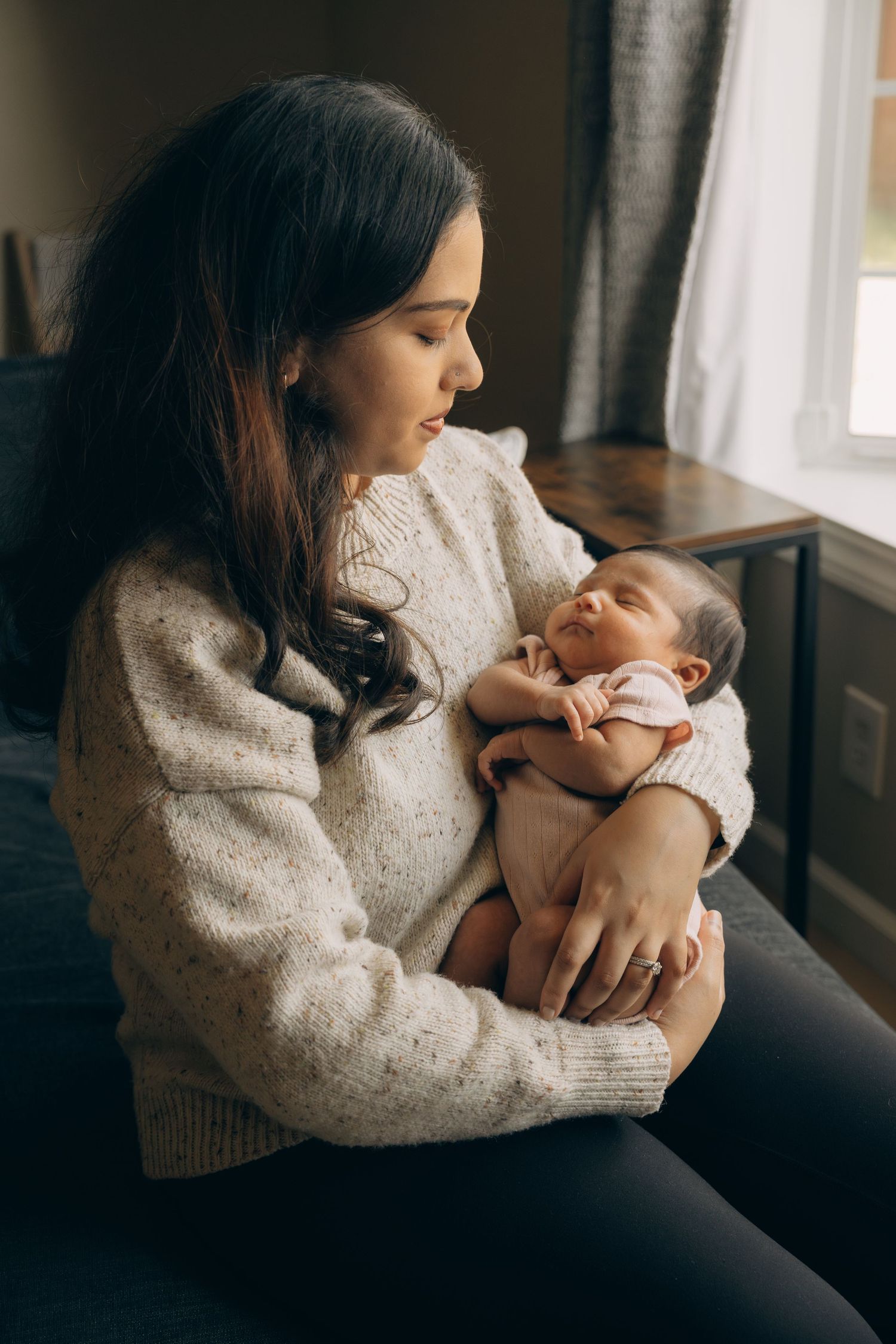 A young mother in a cream sweater tenderly holds her sleeping newborn baby while sitting near a window.