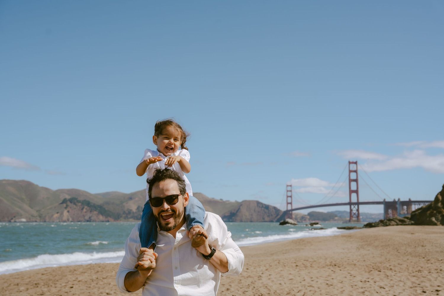 Man with child on shoulders smiling on Baker Beach with Golden Gate Bridge in background on sunny day.