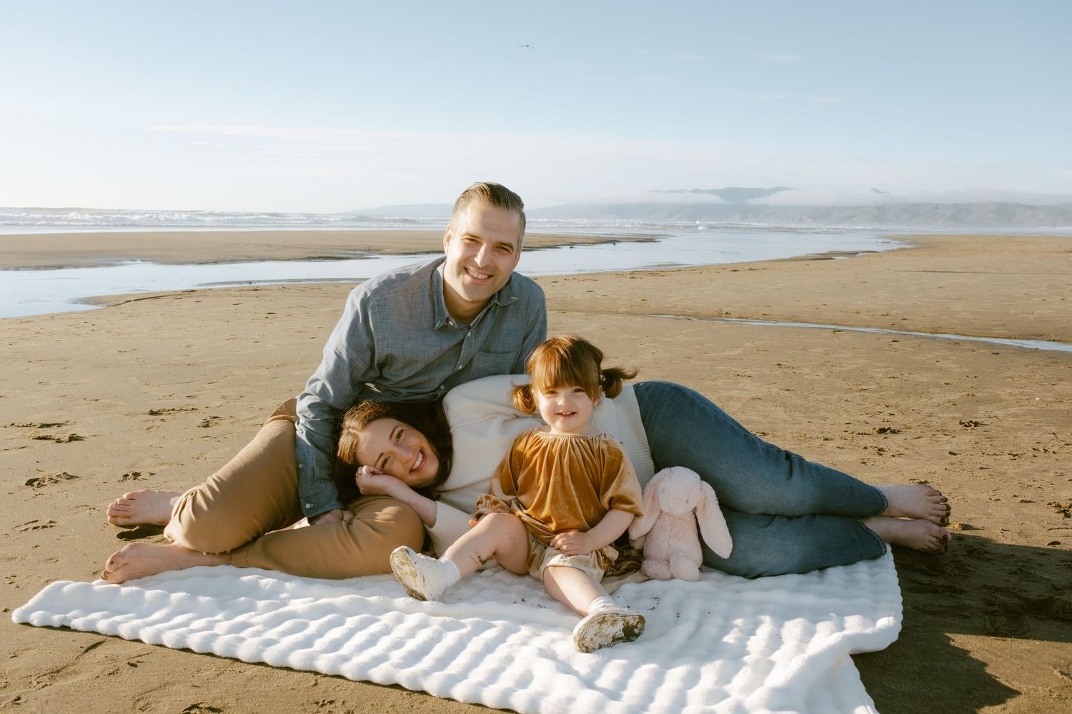 Family of three relaxing on a white blanket on a sandy beach, with ocean waves and clear sky in the background.