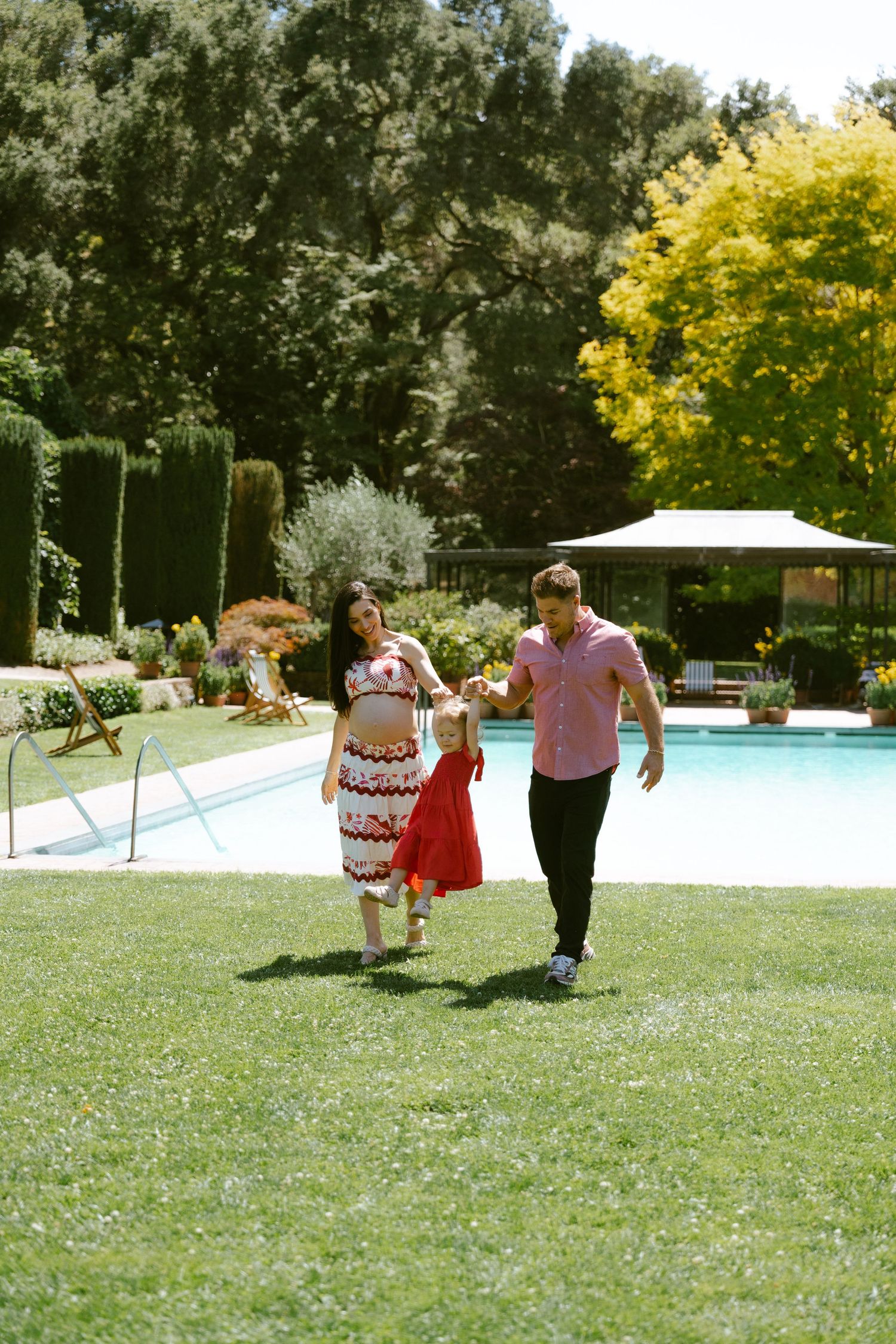 A couple walks hand-in-hand with a child on a sunny lawn beside a pool surrounded by lush greenery.