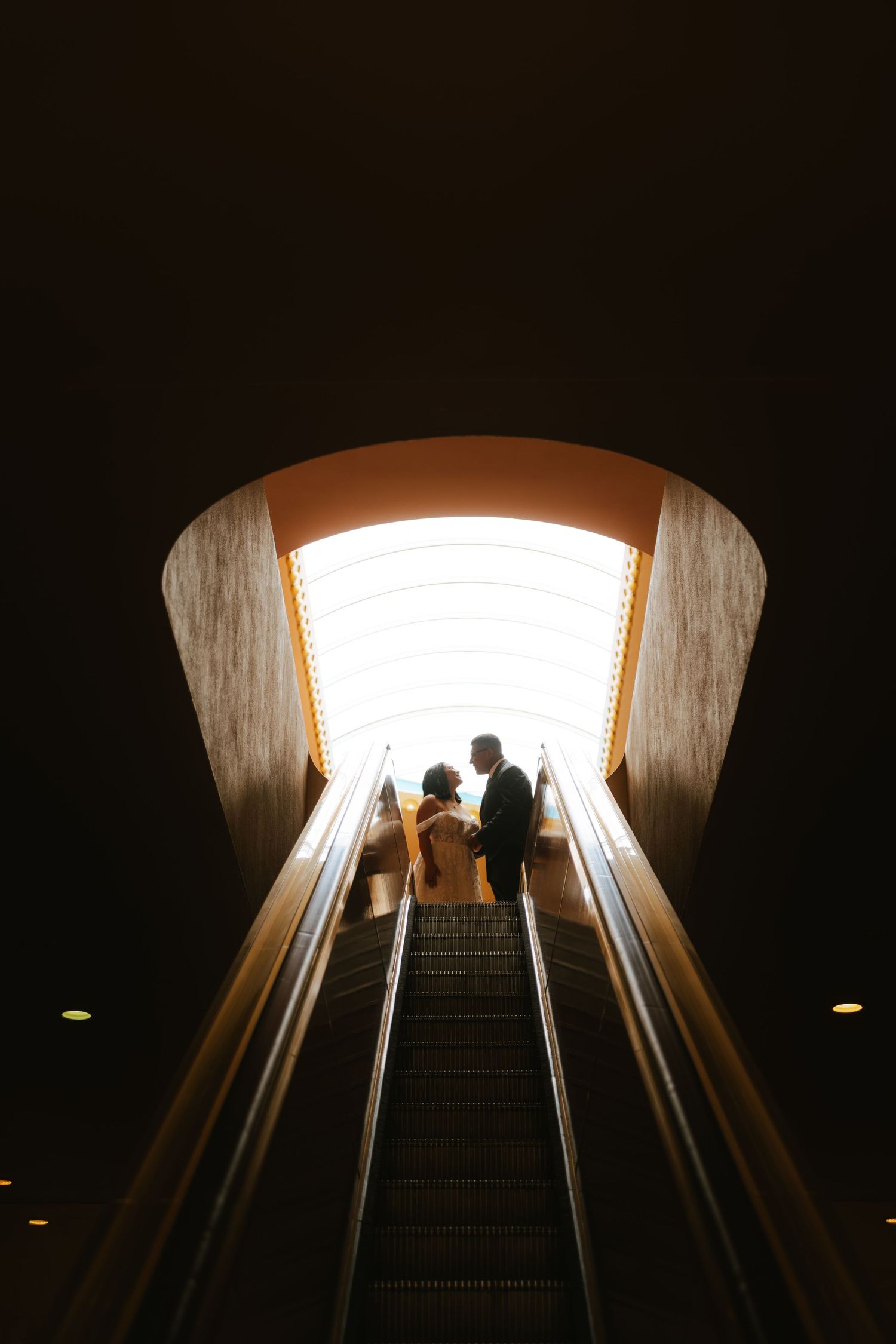 Couple silhouetted on escalator beneath glowing archway in romantic wedding portrait.