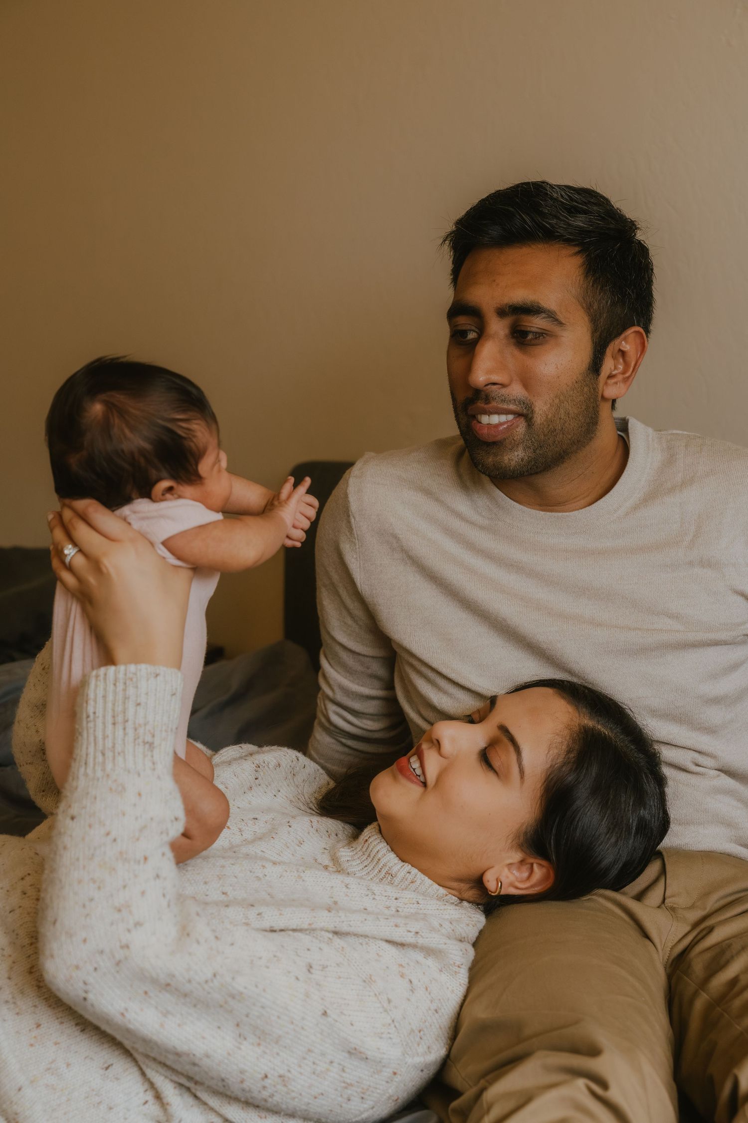 A couple relaxing on a couch, smiling and holding their newborn baby in a warm, cozy home setting.
