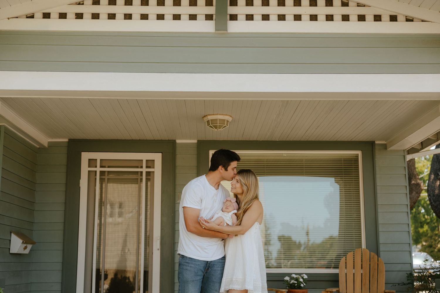 A couple embracing on the front porch of a charming blue house, holding their newborn baby.