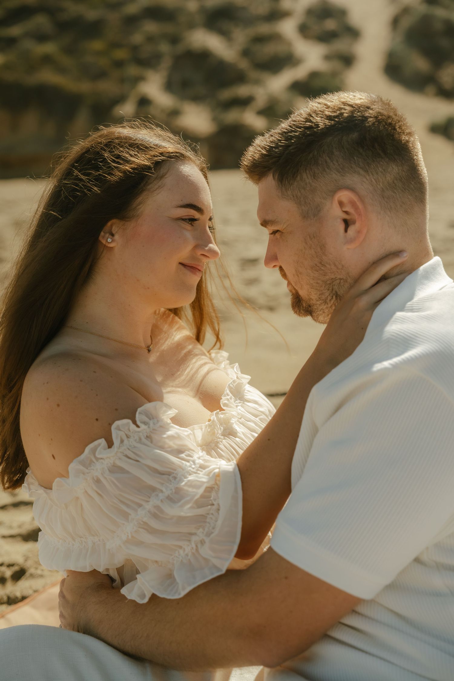 Romantic couple in white outfits sitting close together on sandy beach gazing into each others eyes.