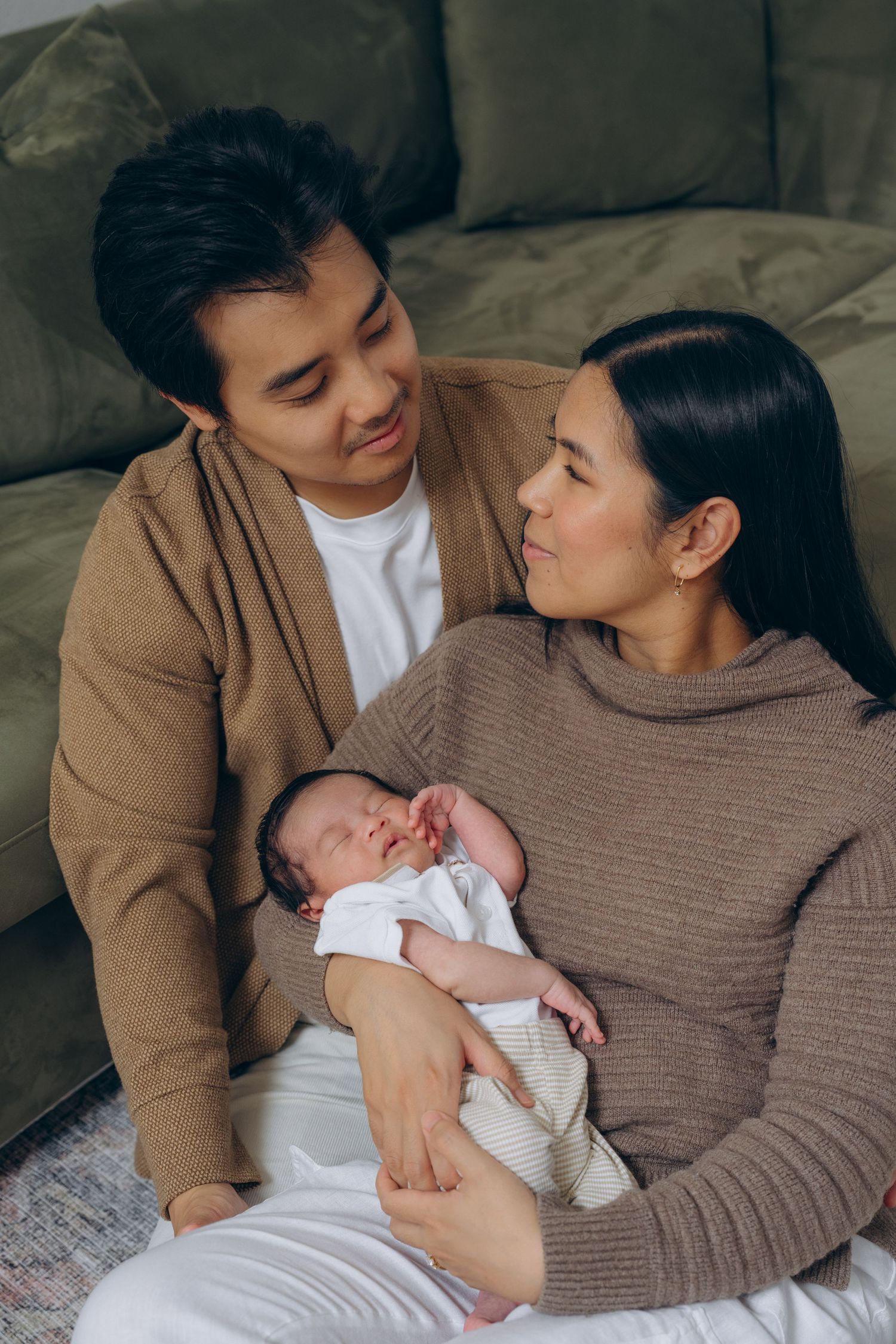 Asian couple sitting on floor holding their newborn baby, sharing a tender moment together at home.