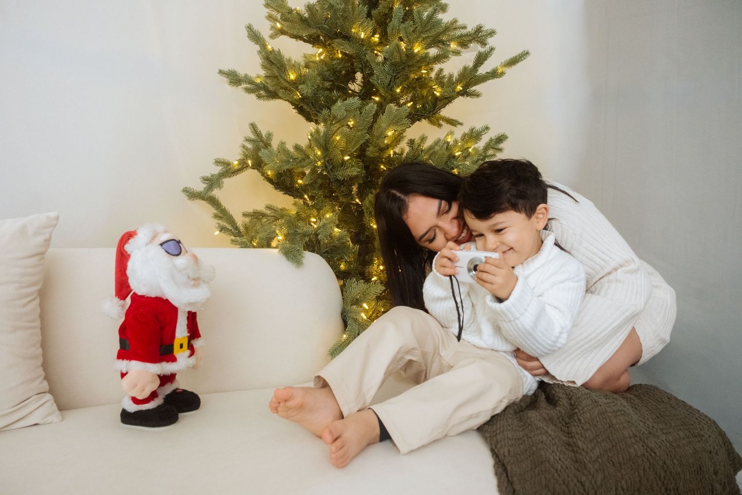 A cozy holiday scene with people lounging on a white couch next to a lit Christmas tree and Santa decoration.
