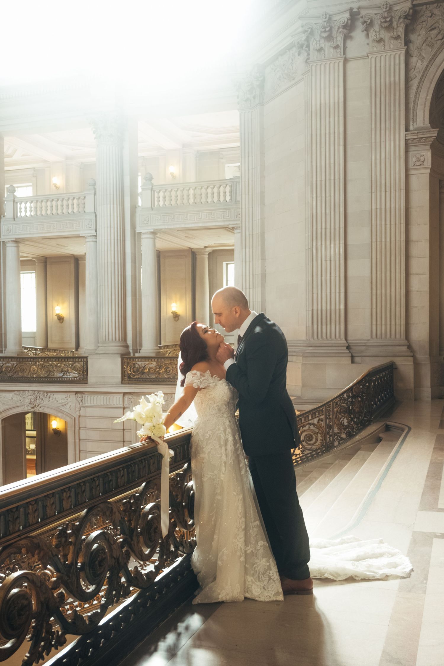 A romantic wedding couple dances in an elegant marble hall with ornate railings and classical columns bathed in soft light.
